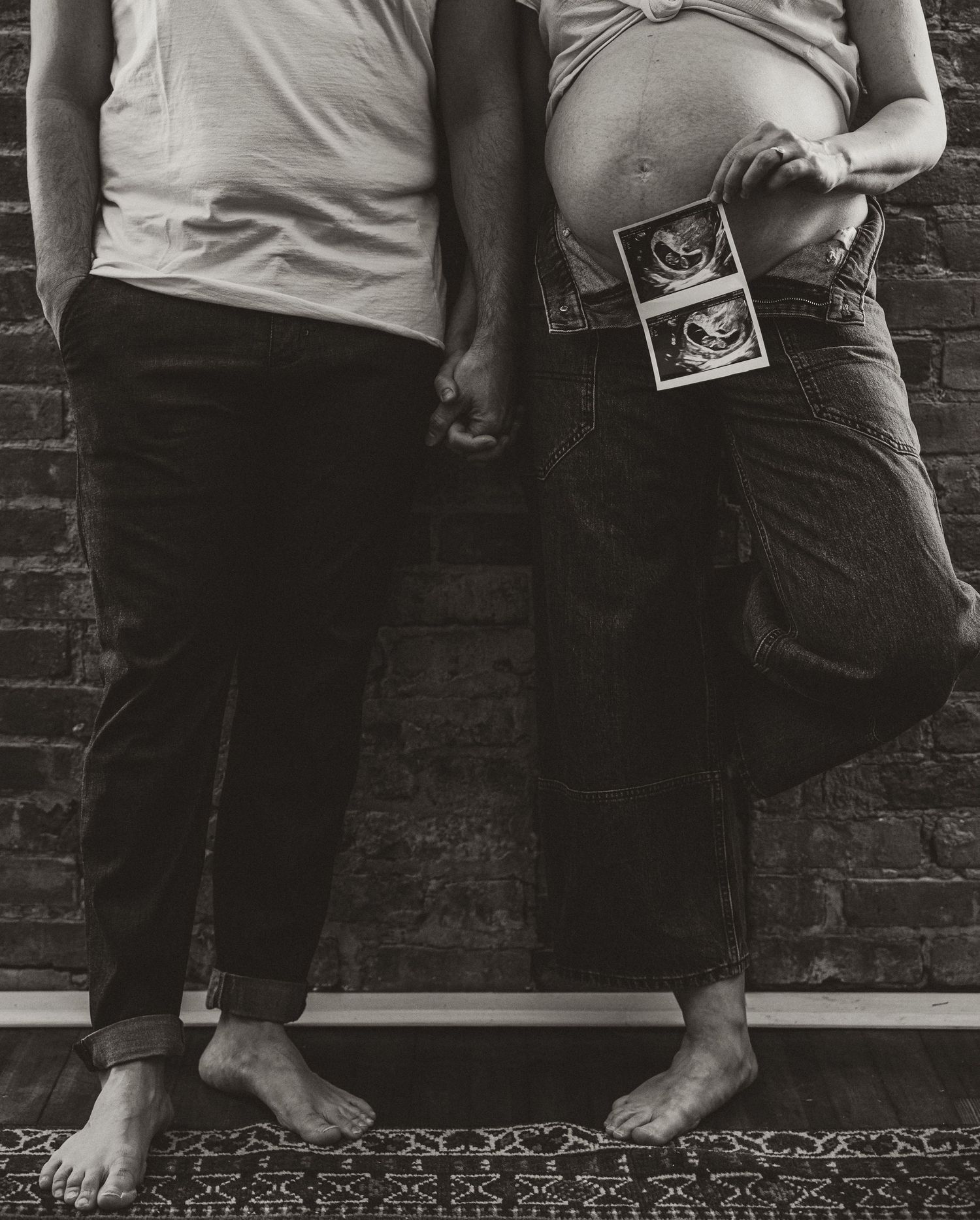Black and white photo of couple's legs with ultrasound photos against brick wall.