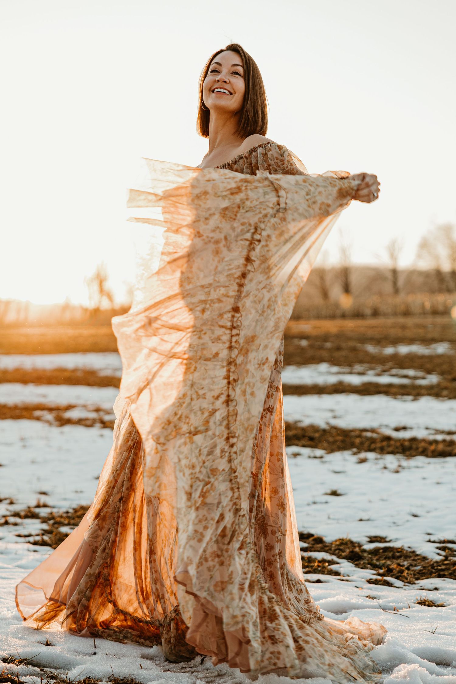 A person in a flowing lace dress poses gracefully in the snow at sunset creating an ethereal winter scene.