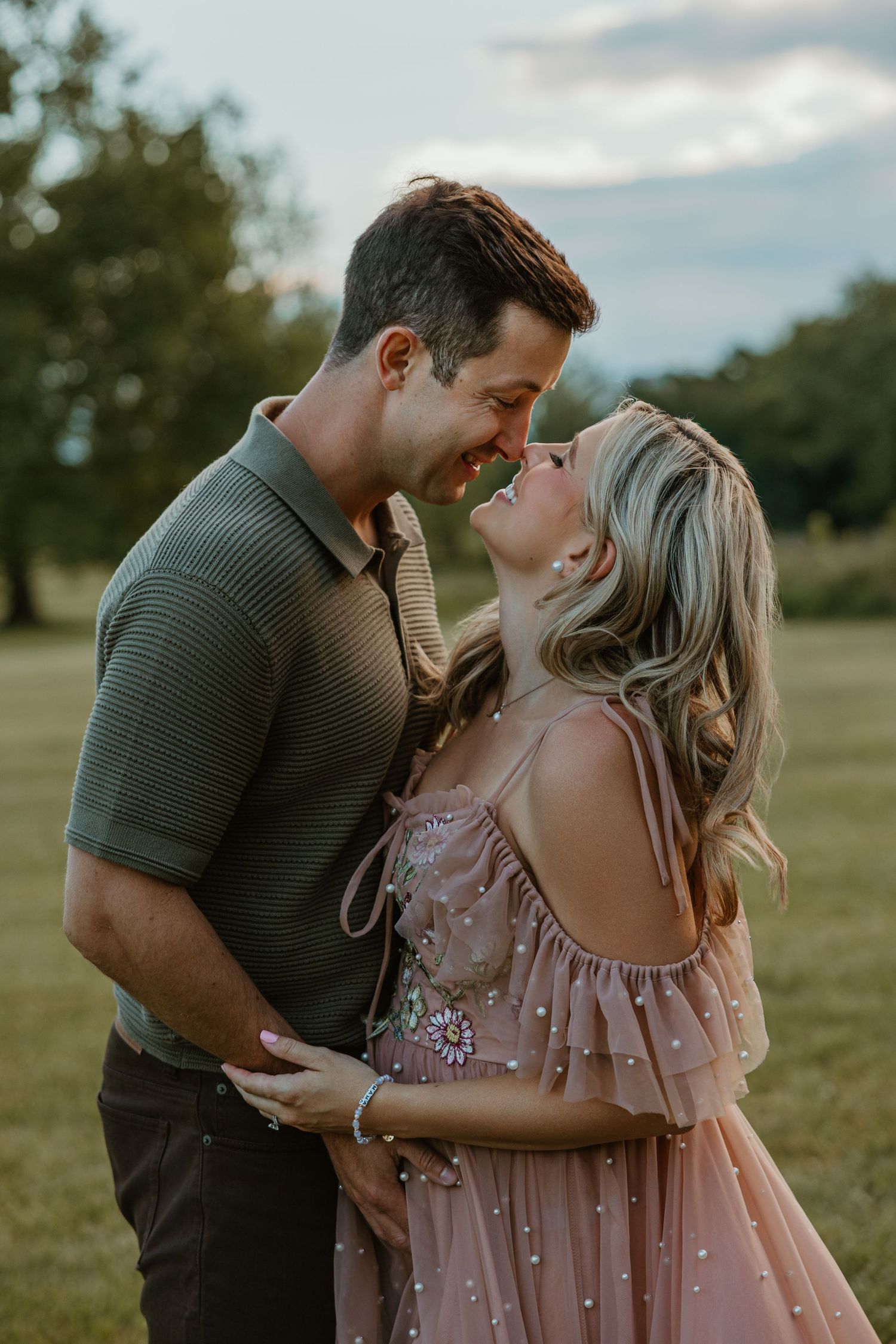 A couple shares a romantic moment outdoors, smiling at each other in a lush green field at golden hour.
