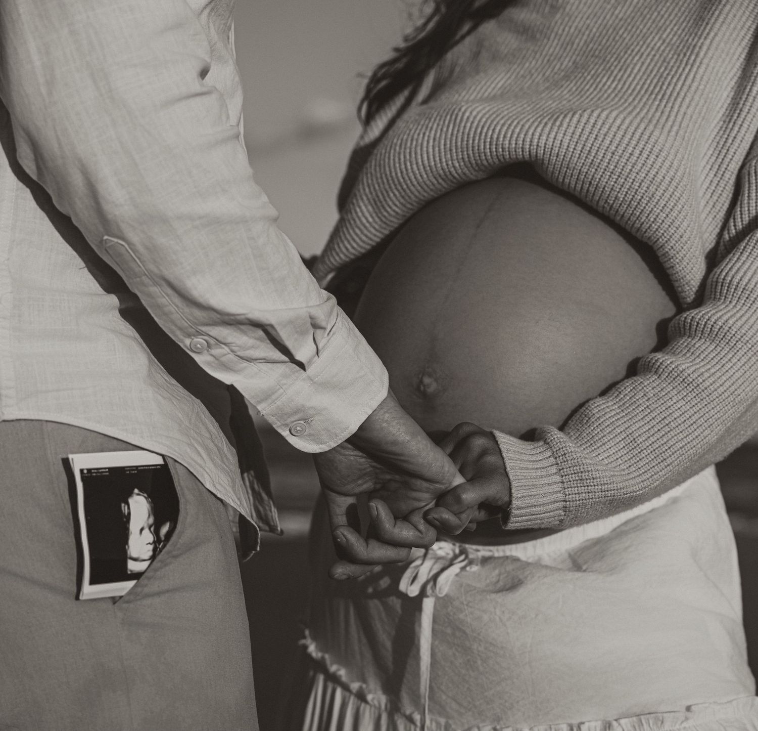 Close-up black and white maternity photo of couple holding hands with ultrasound photo tucked in pocket.
