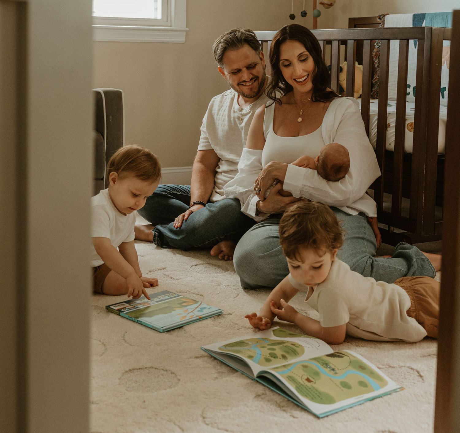 Family reads books together on nursery floor, mother holding newborn as children enjoy storytime.