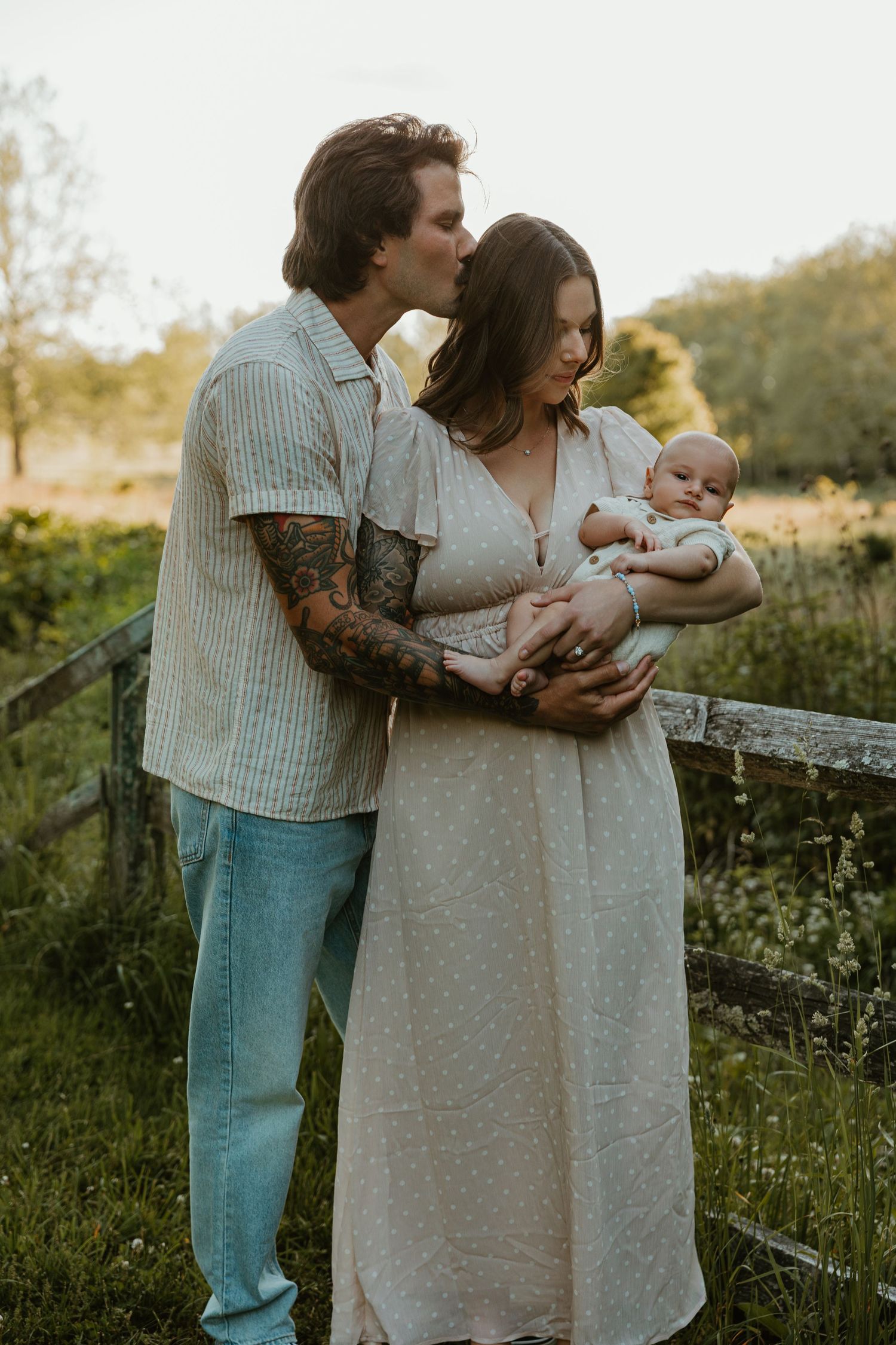 A tattooed couple shares a tender kiss while cradling their newborn baby outdoors at golden hour.