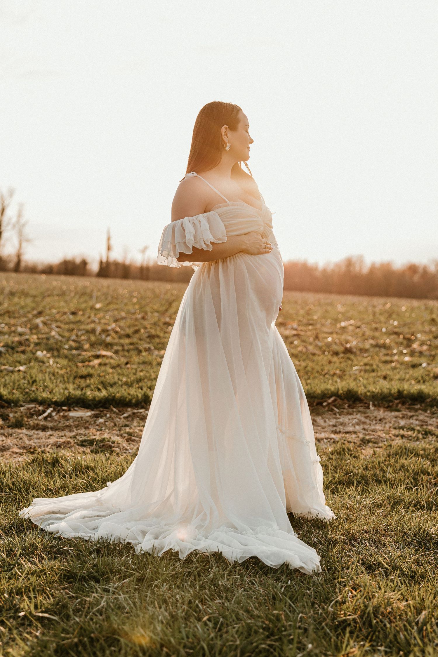 Pregnant woman in flowing white off-shoulder gown standing in field at golden hour sunset.