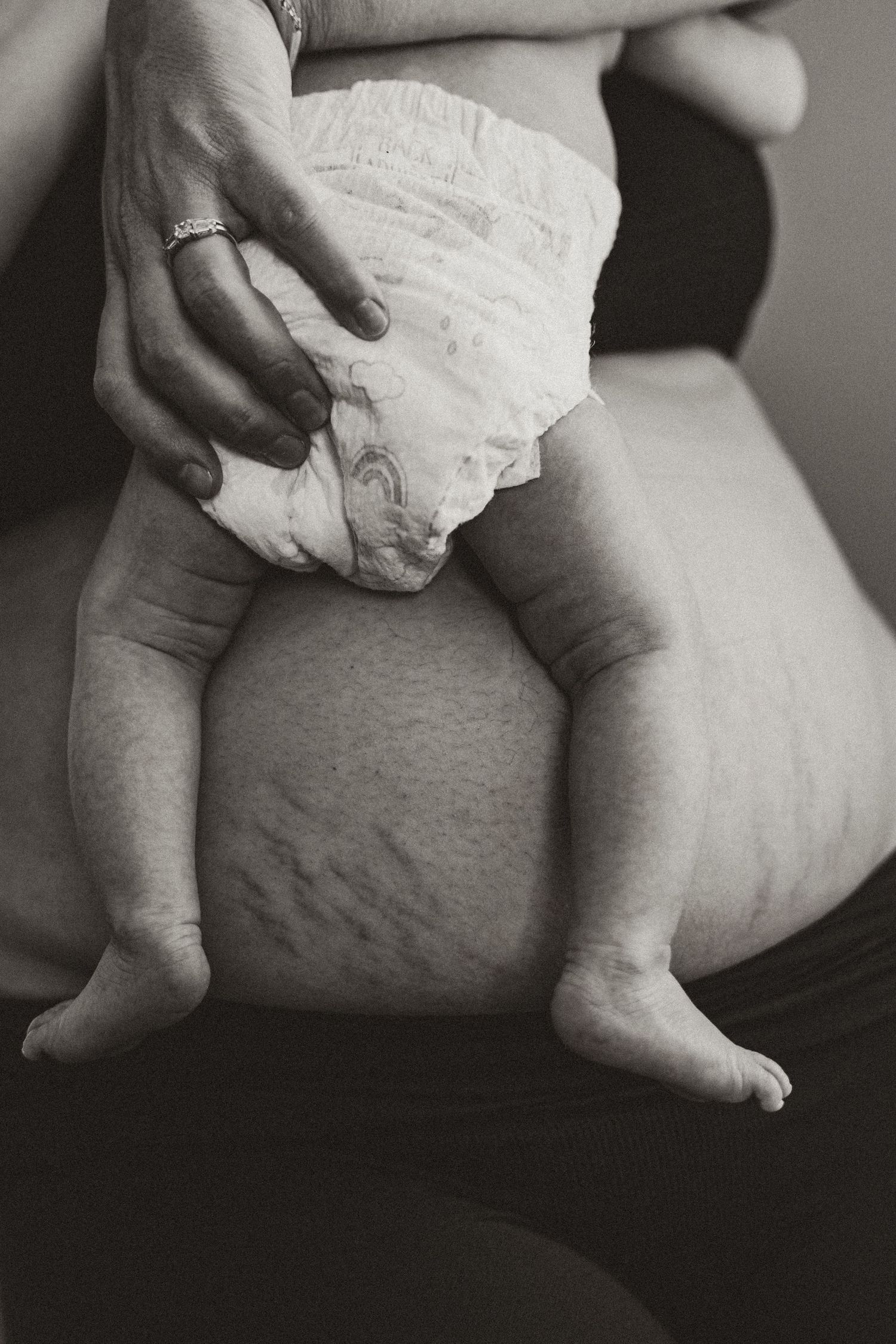 Black and white photo of a newborn baby in a diaper sitting on a mothers lap showing postpartum stretch marks.
