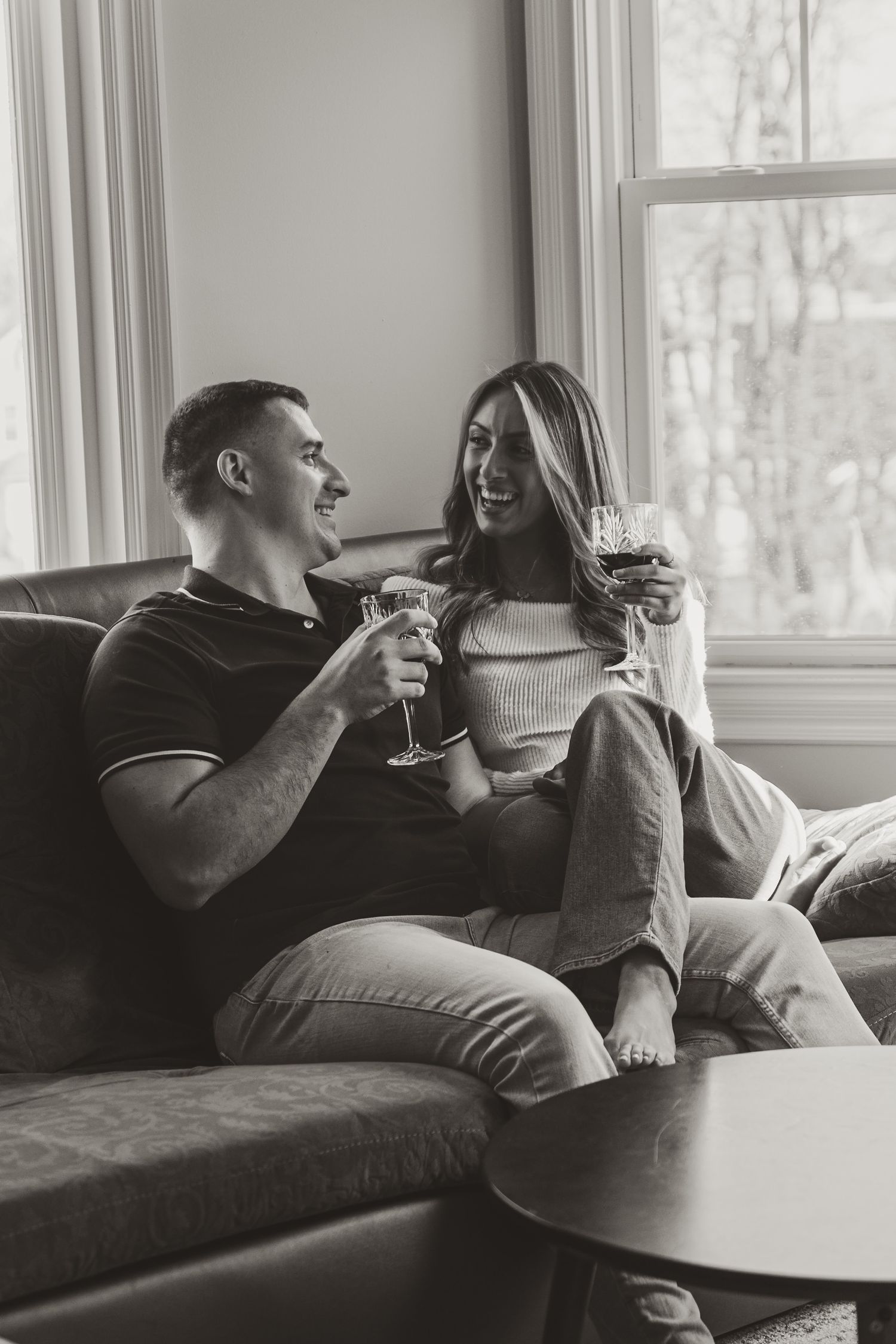 A couple relaxing on a couch by a window sharing wine and intimate conversation.