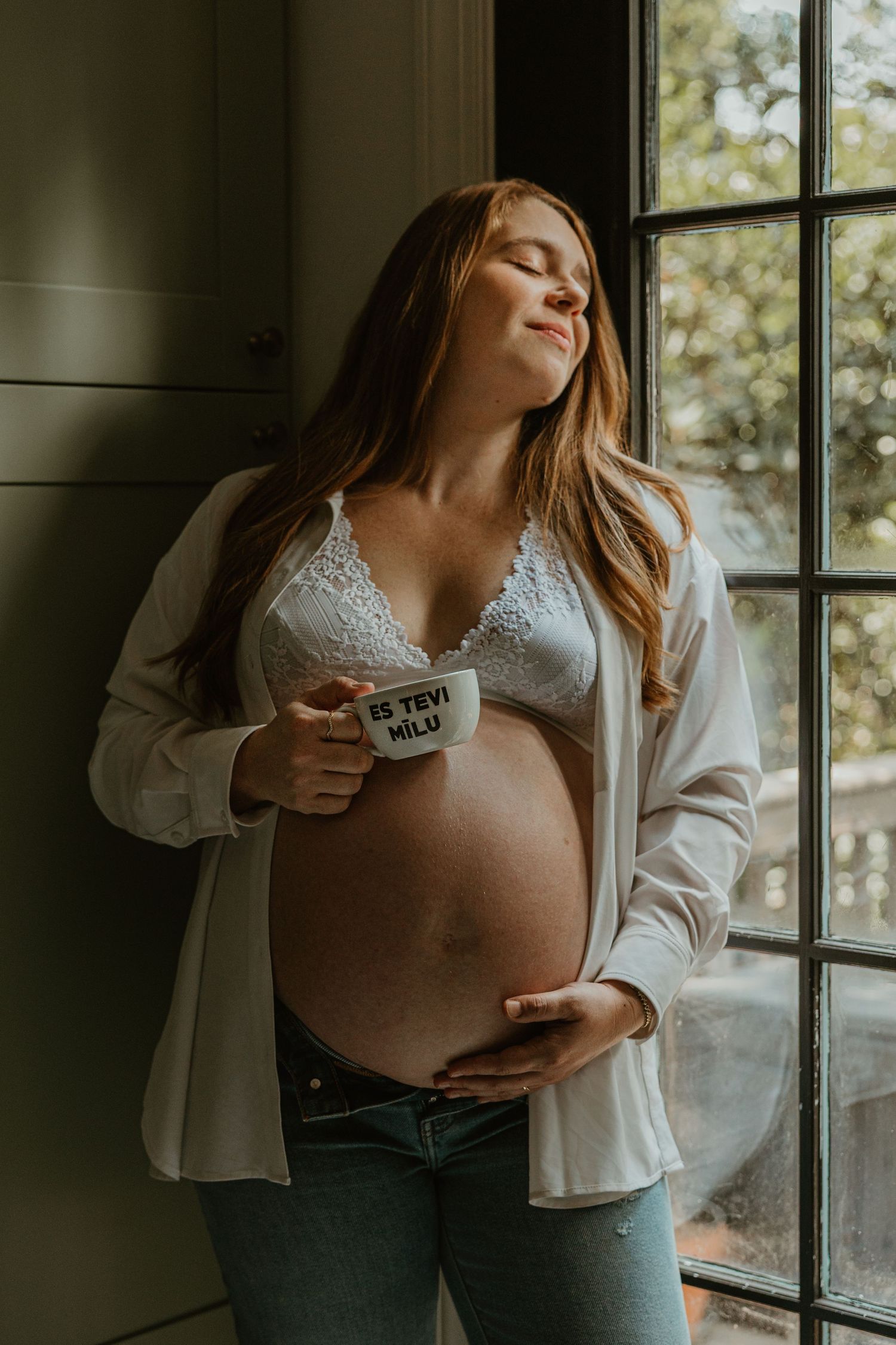 Pregnant woman in white lace bra and open shirt holds mug near window, cradling baby bump with eyes closed.