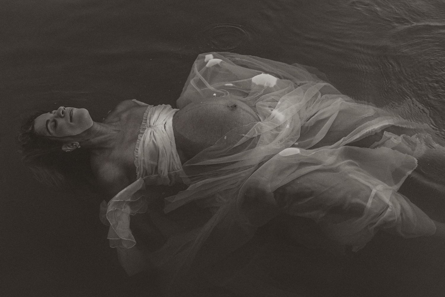 Black and white photo of a pregnant woman floating in water wearing a sheer white flowing gown.