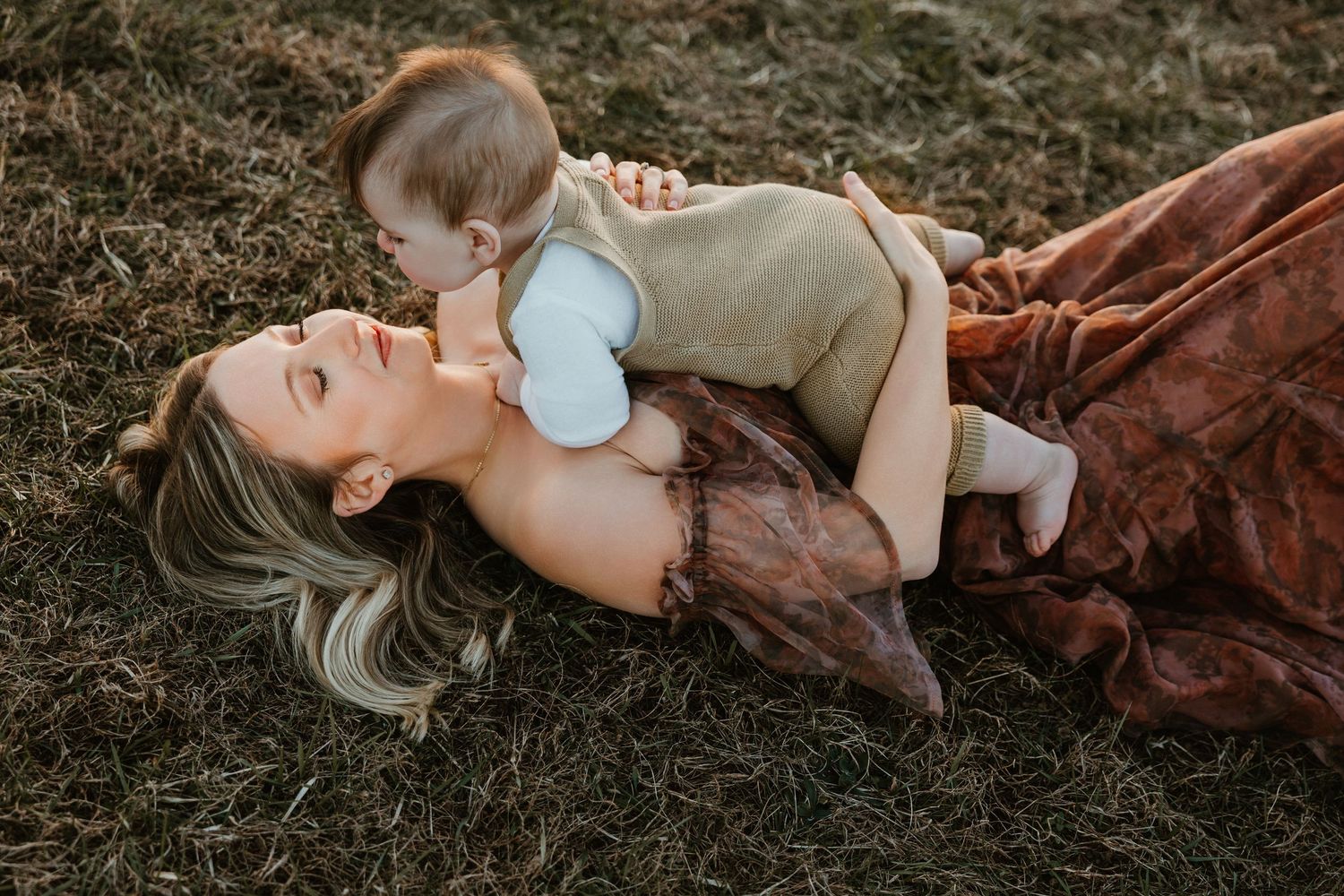 Mother lying on grass holding baby above her, sharing a tender moment outdoors in a rustic brown dress.
