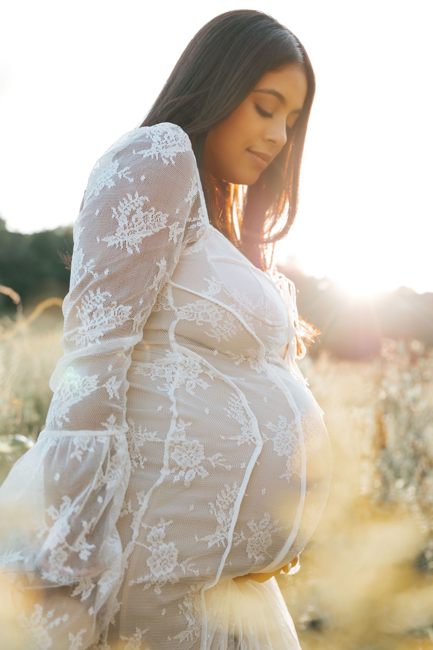 A woman in a white lace dress poses in a sunlit field during golden hour.