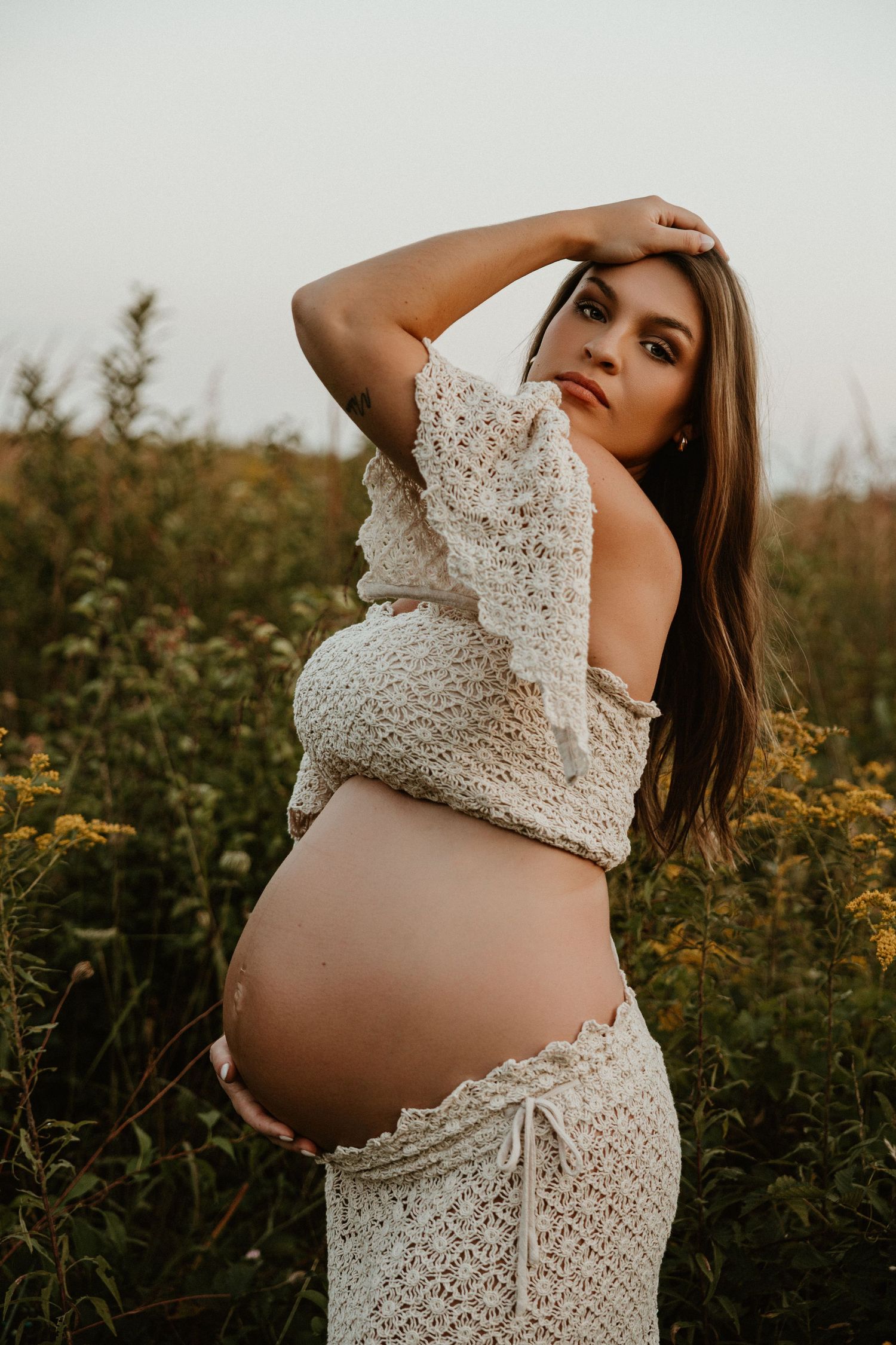 Artistic maternity portrait in a wildflower field at sunset featuring white lace attire.
