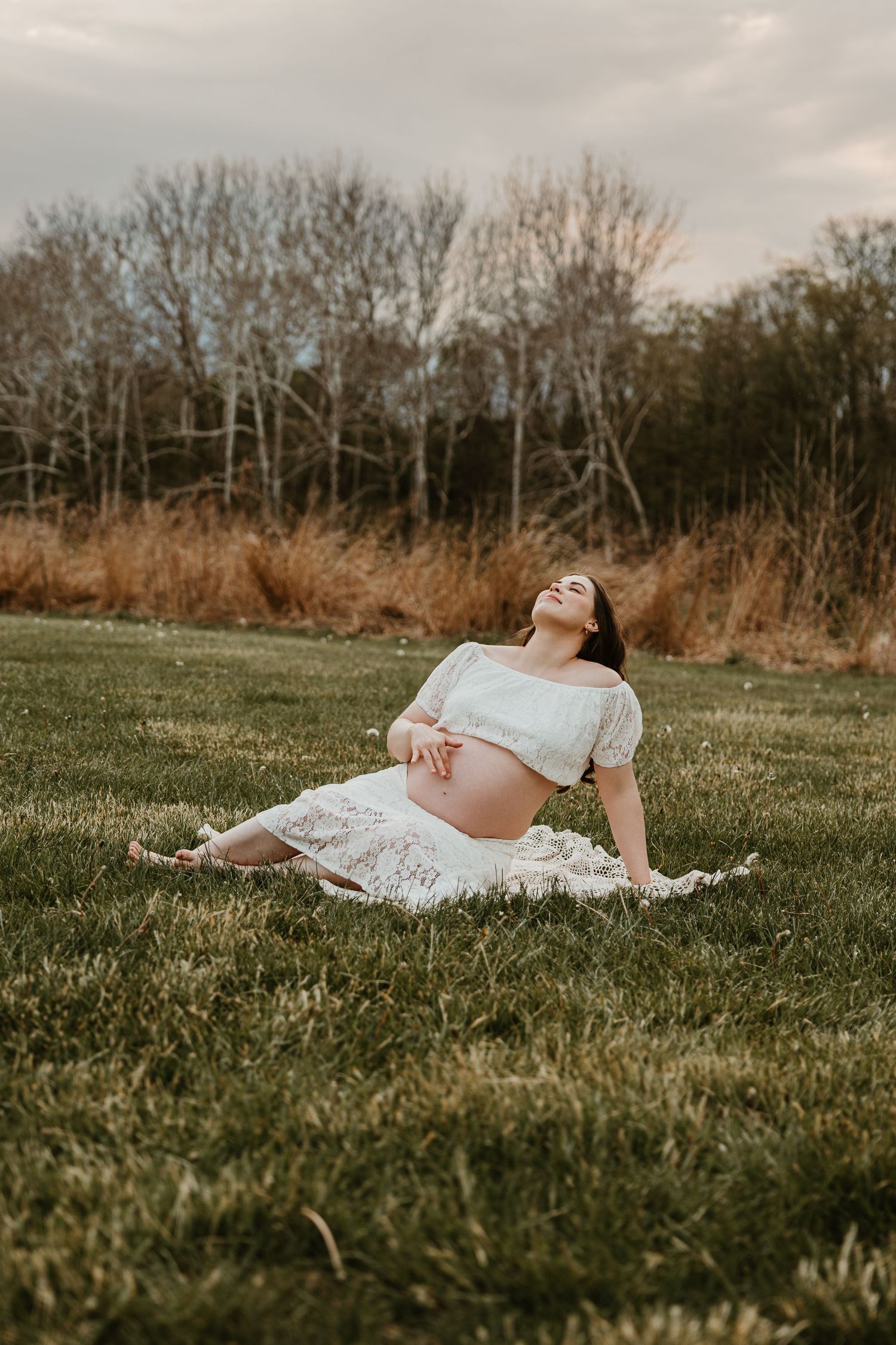 Pregnant woman in white crop top and skirt sitting on grass in a field, smiling with head tilted back.