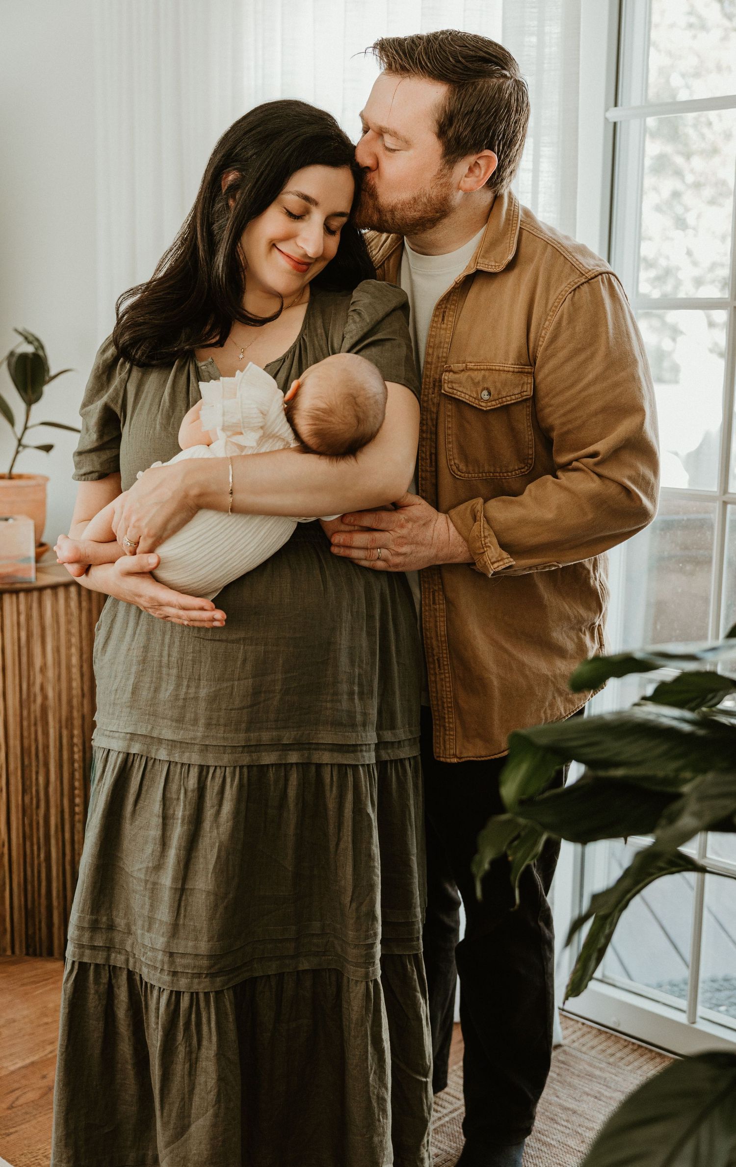 A couple tenderly holds their newborn baby, the man kissing the woman's head, in a bright, plant-filled room.