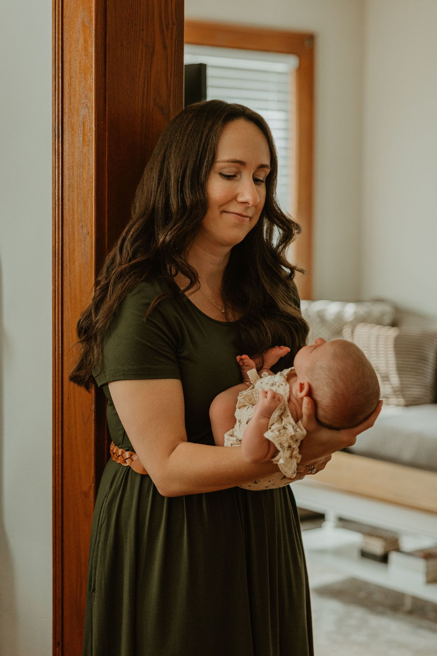 A woman in an olive green dress tenderly holds a newborn baby while leaning against a wooden door frame.