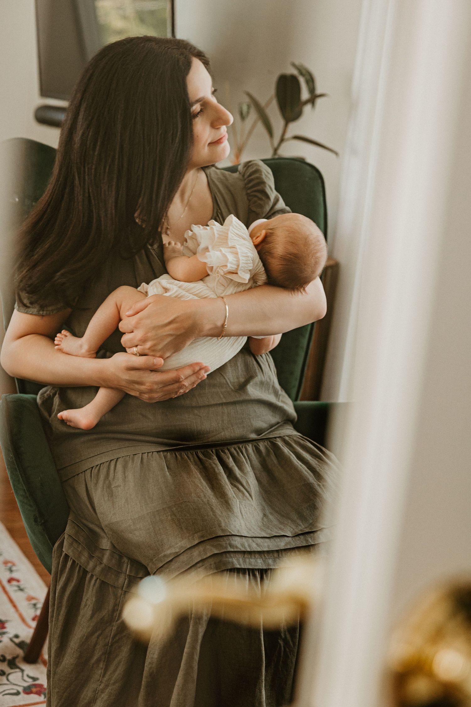 A mother in an olive dress sits in a green chair, cradling her newborn baby near a sunlit window.