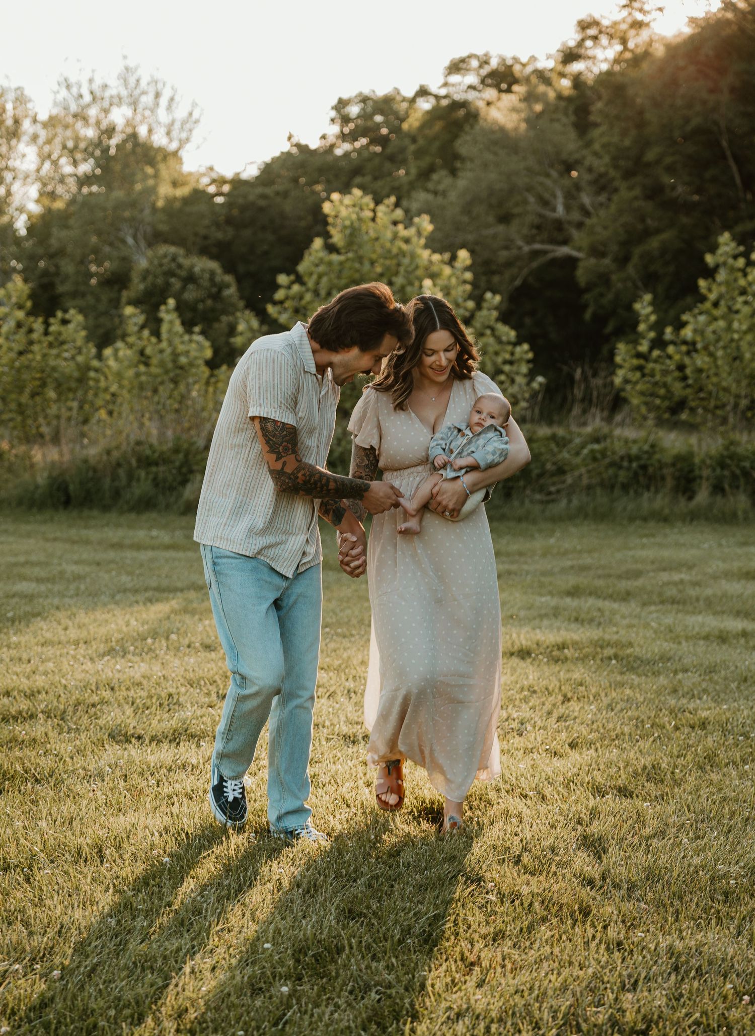 A young family with tattoos walks through a sunlit meadow, parents smiling at their newborn baby.