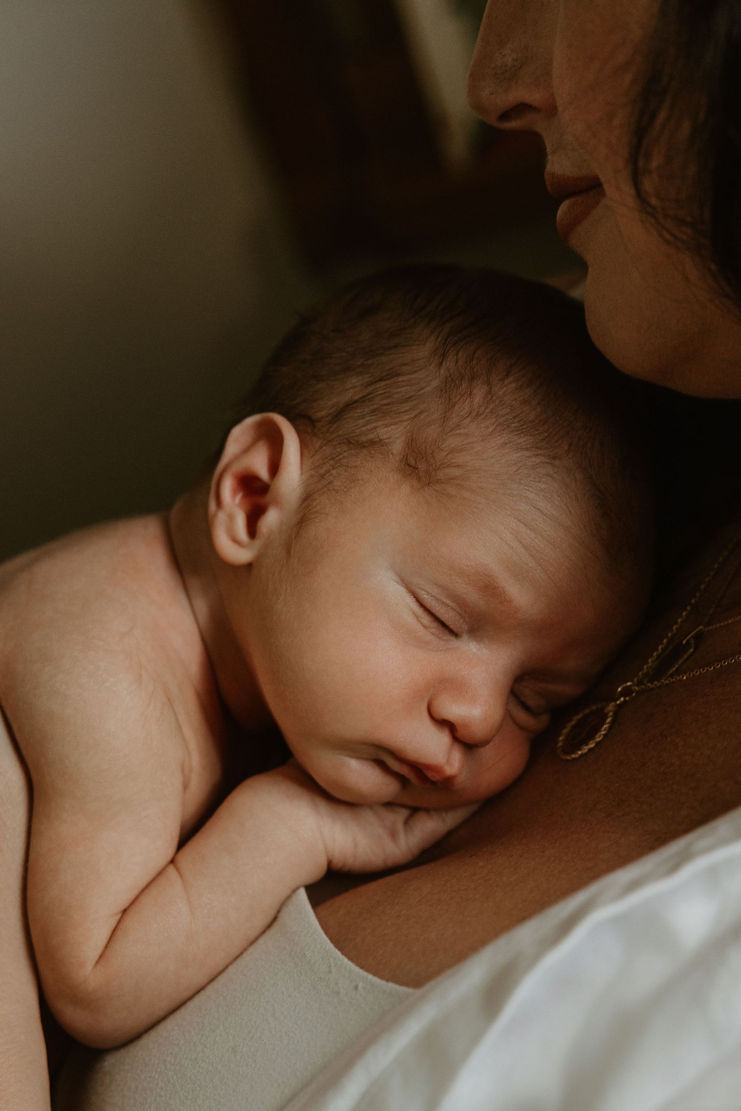 A peaceful newborn baby sleeping on mother's chest in a warm, intimate close-up portrait photo.