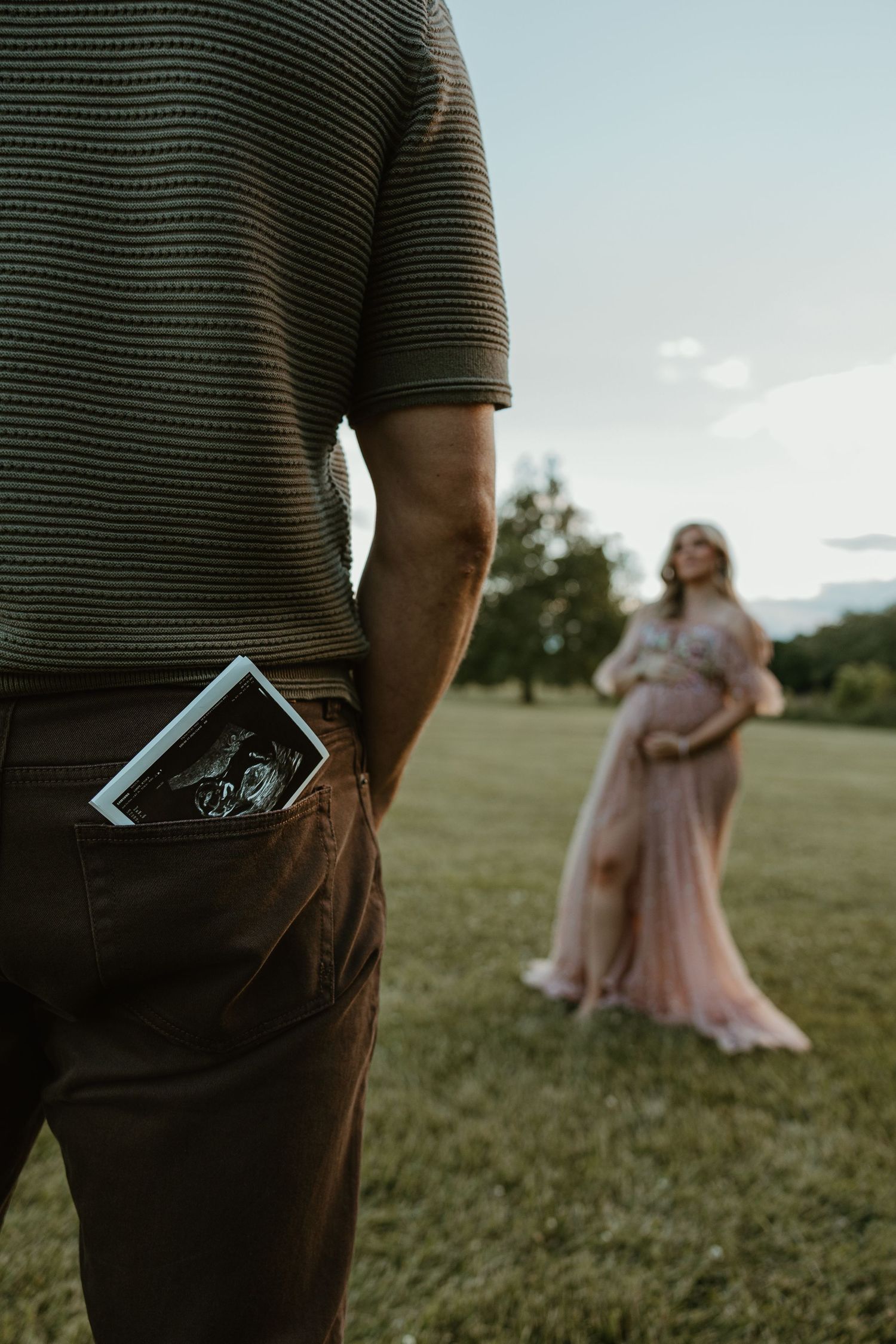 Man with ultrasound photo in pocket stands before pregnant woman in pink dress during outdoor maternity photoshoot.