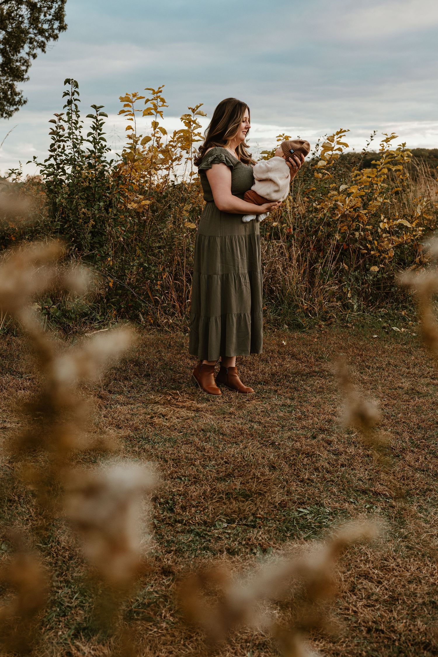Person in olive dress standing among autumn foliage at sunset.