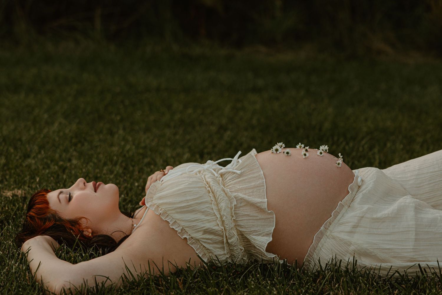 Pregnant woman in white dress lying on green grass, serene maternity photo shoot outdoors.