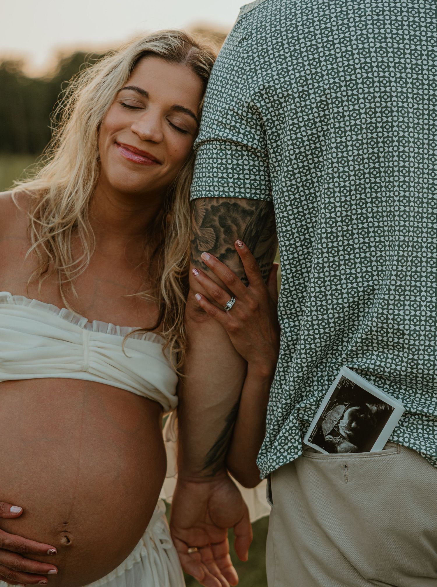 Pregnant woman in white dress leans on partner holding ultrasound photo during outdoor maternity photoshoot at sunset.