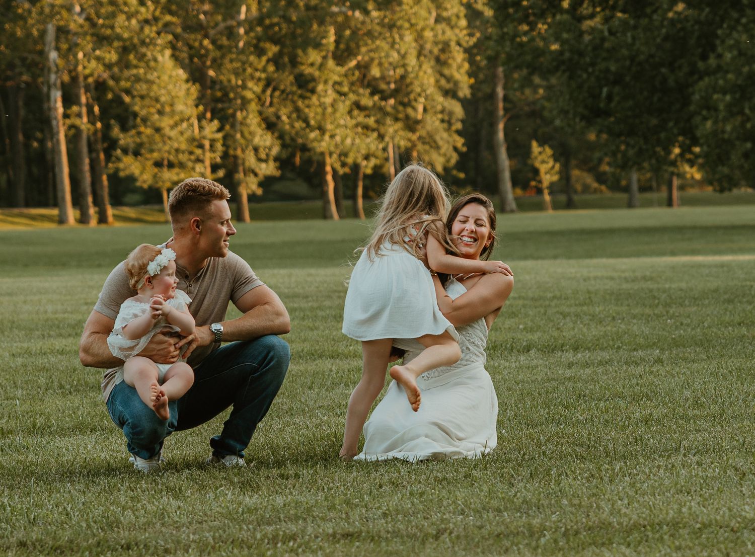 Family portrait on green lawn with father holding baby, mother hugging young child, trees in background at golden hour.