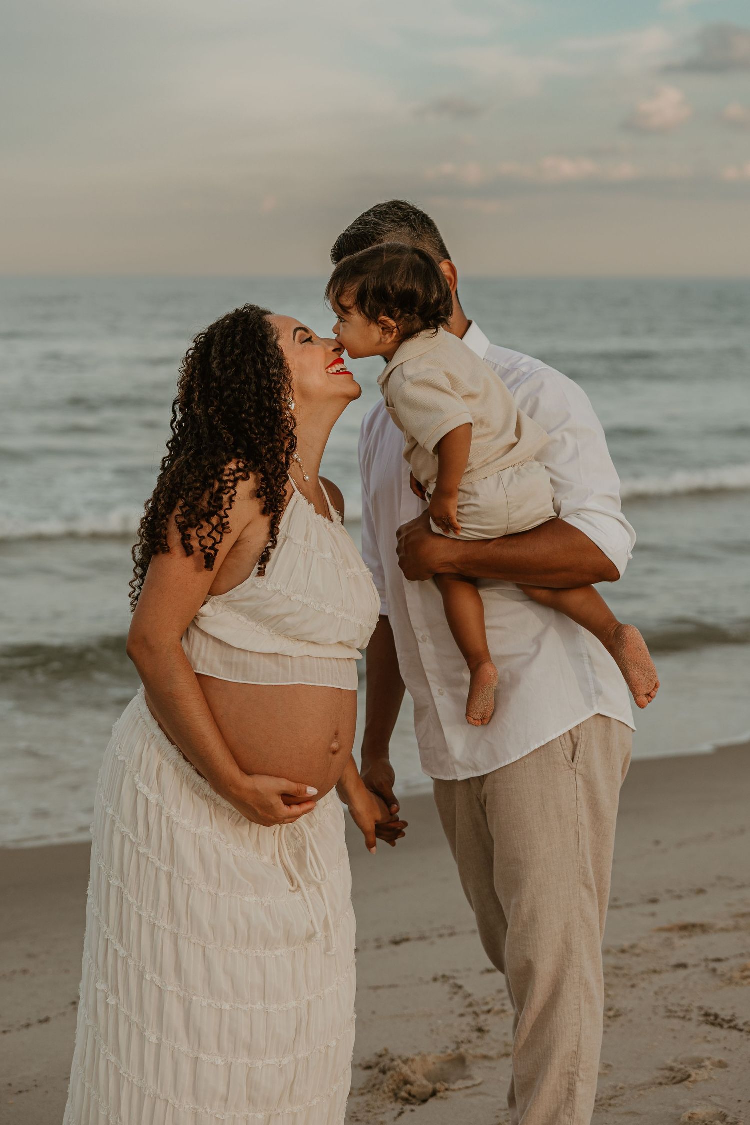 Pregnant woman in white dress with partner holding toddler sharing a tender moment on a beach at sunset.