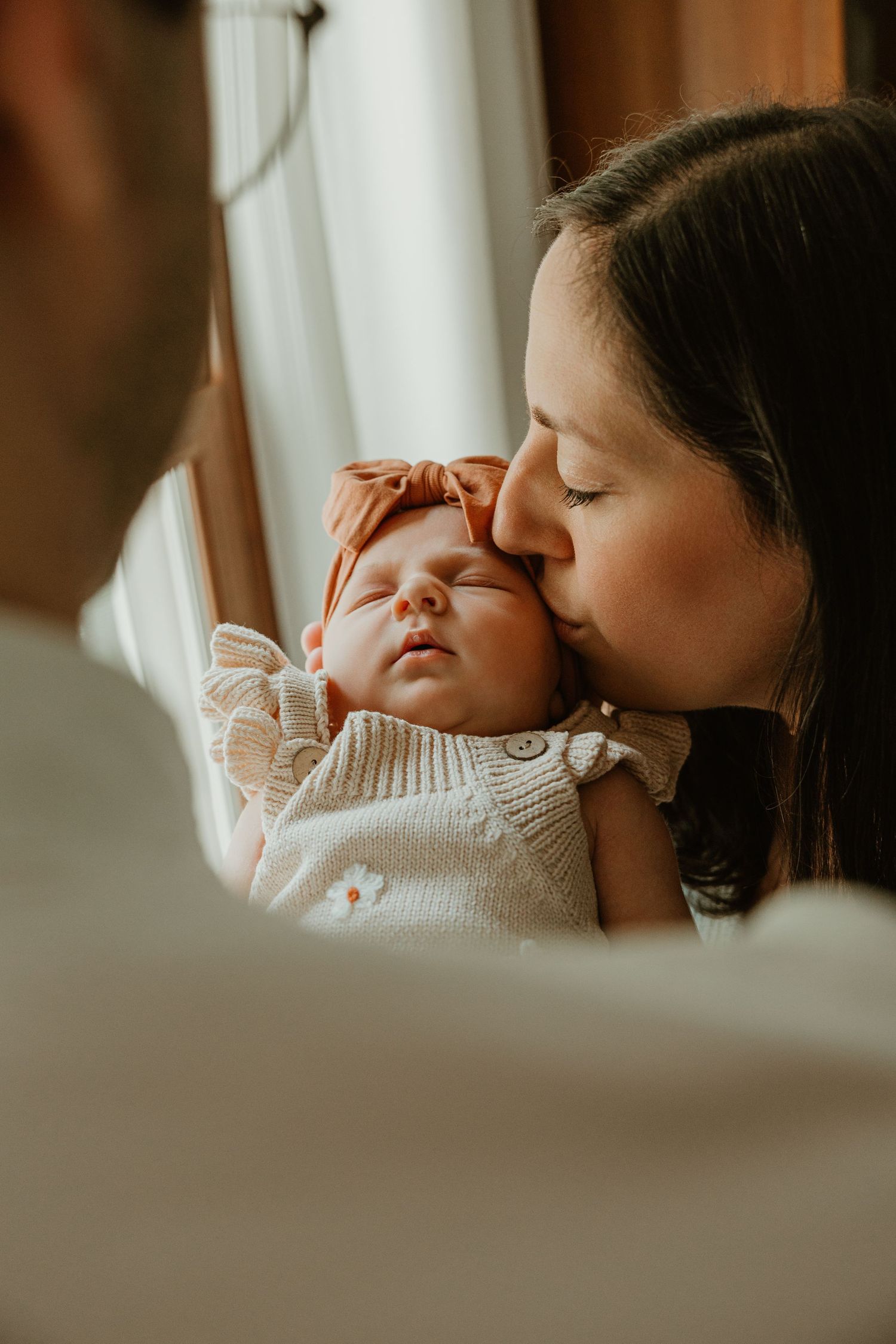 A mother tenderly kisses her sleeping newborn baby wearing a rust bow headband and cream knit outfit.