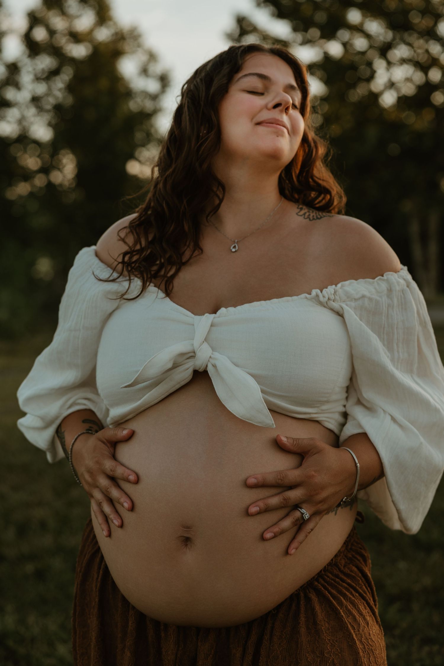 Pregnant woman in white off-shoulder crop top holds baby bump outdoors at golden hour.