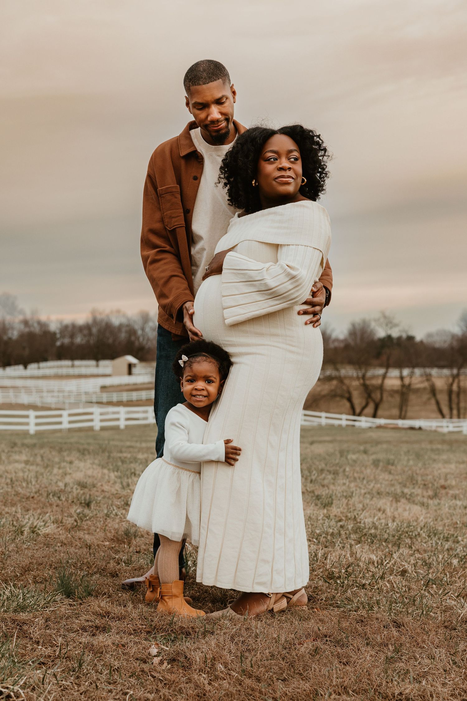 A pregnant Black woman in a white dress poses with her partner and toddler daughter on a farm at dusk.