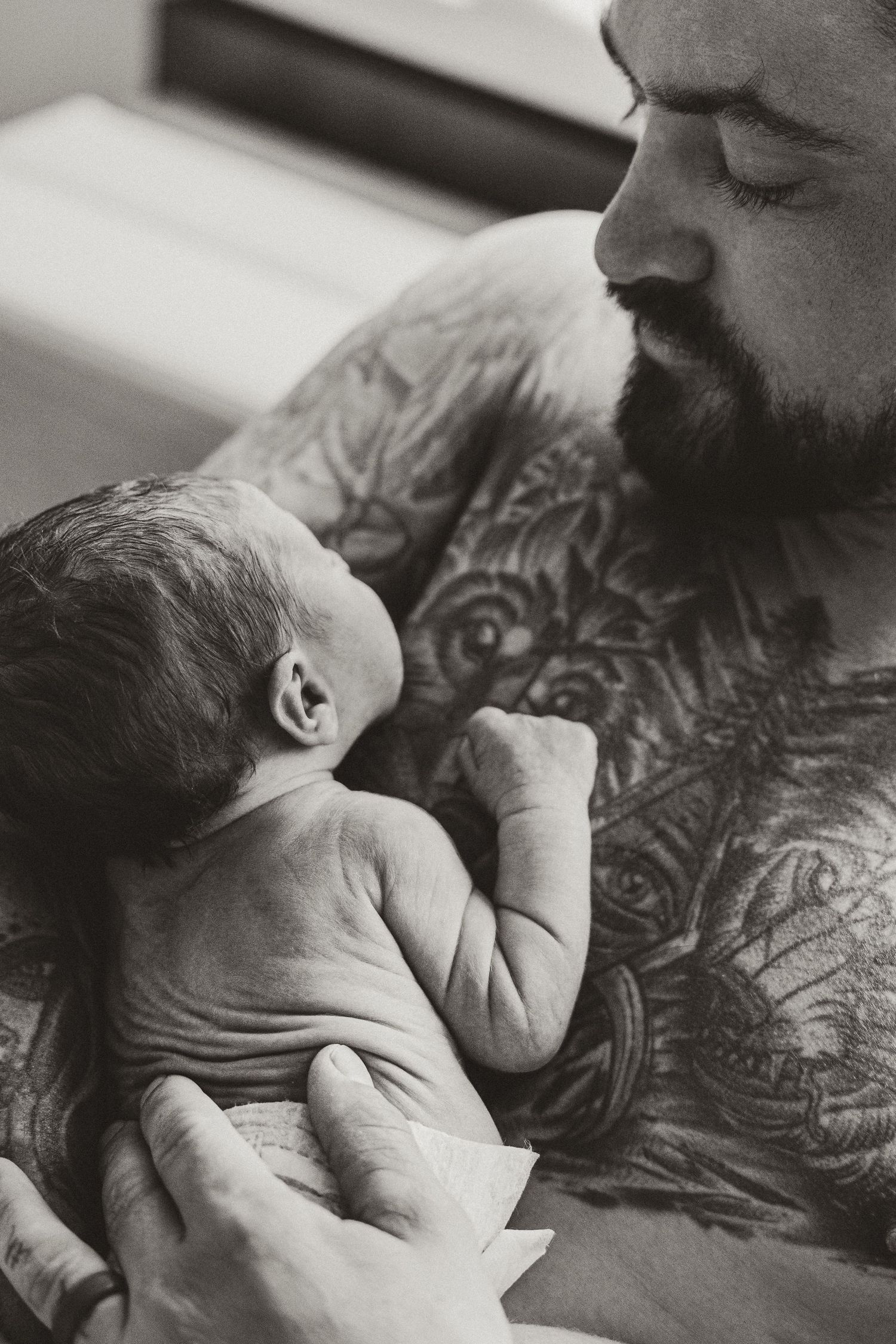 Tattooed father tenderly holding his newborn baby skin-to-skin in black and white photo.