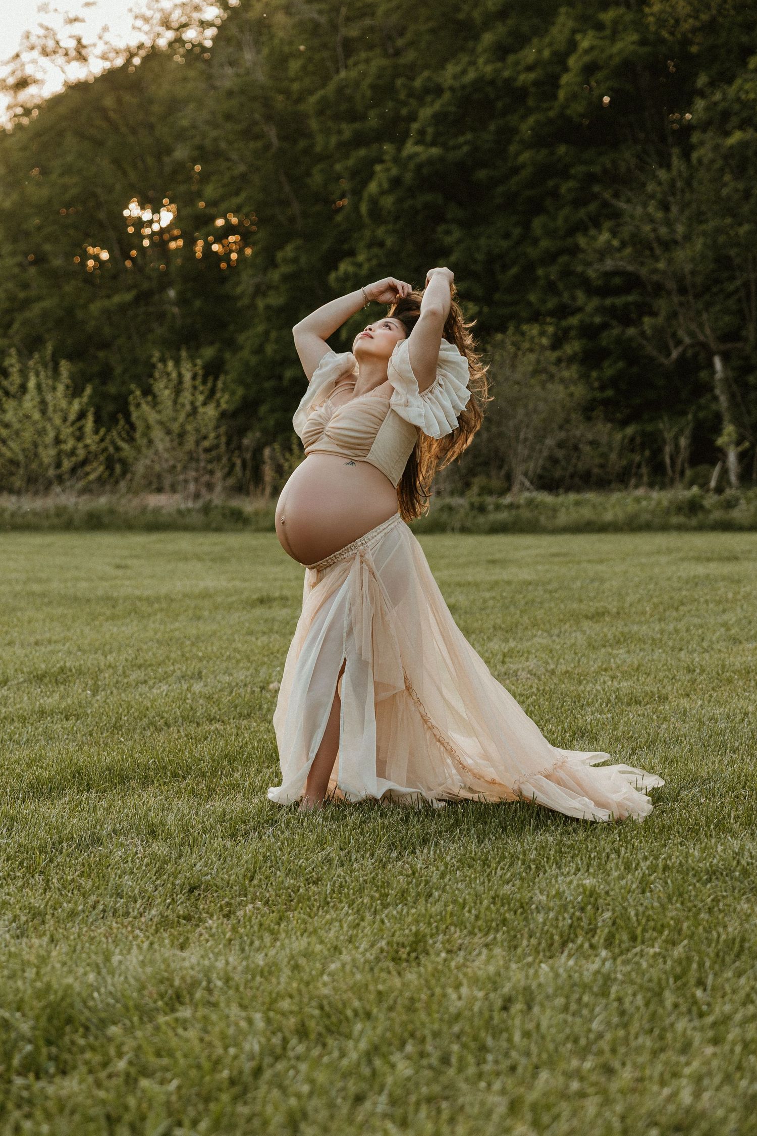 Pregnant woman in elegant beige maternity gown posing gracefully in lush green field at golden hour sunset.