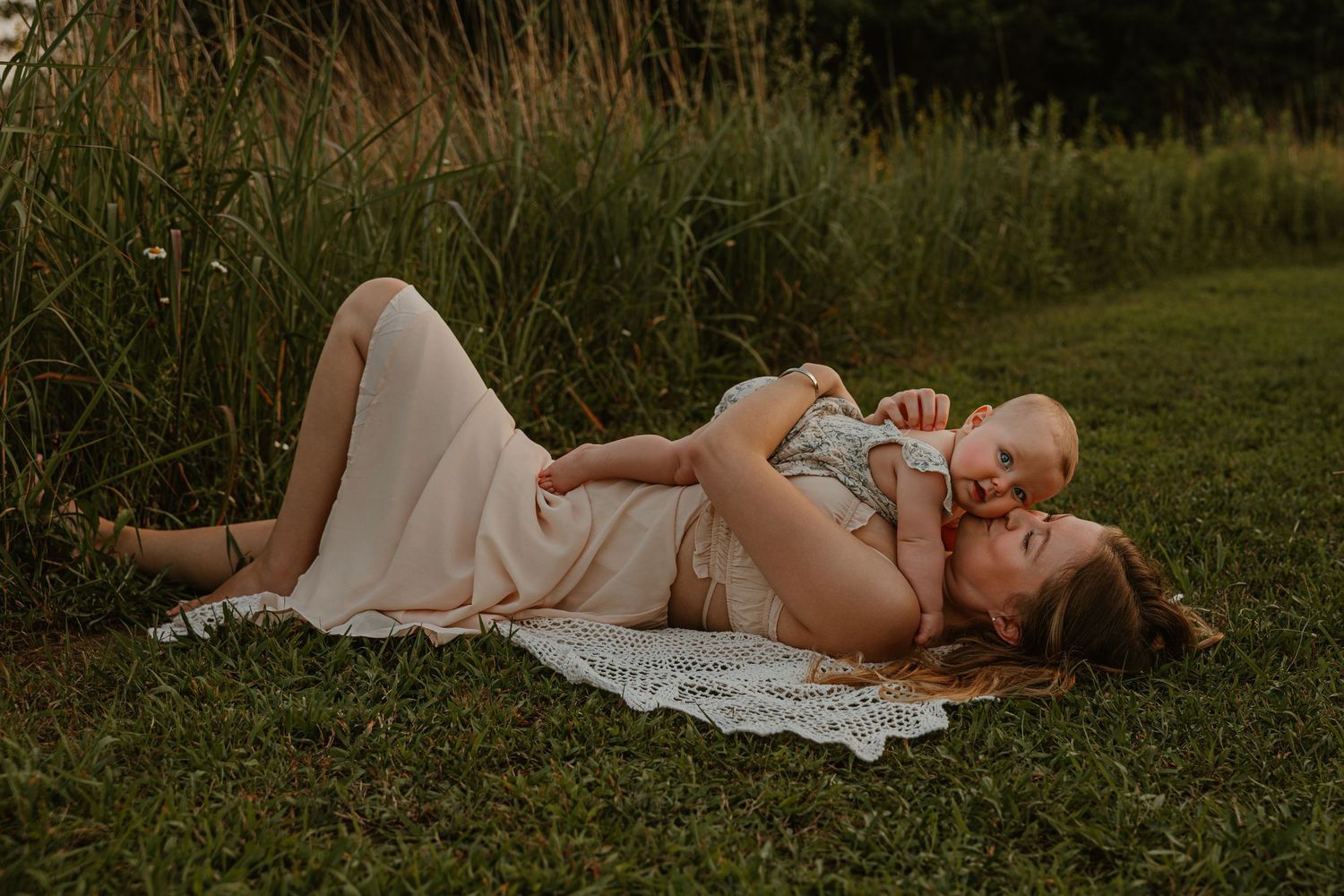 A mother lies on a blanket in grass, lovingly holding her laughing baby up in the air during a golden hour outdoor session.