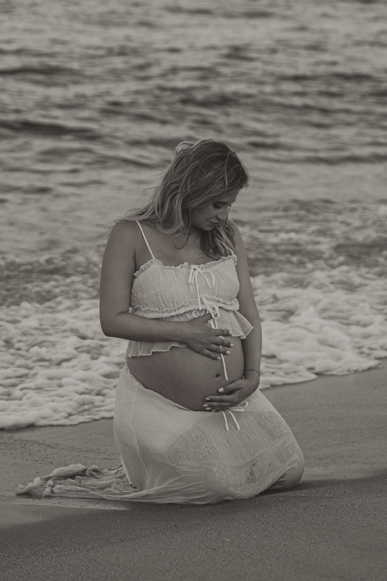 Pregnant woman kneeling on beach shore in white dress, cradling baby bump, black and white maternity photo.