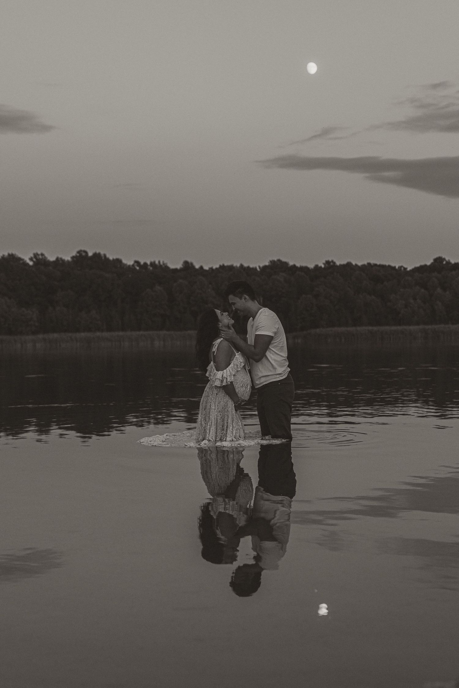 Romantic couple embracing in a moonlit lake at dusk, with their reflection mirrored on the calm water surface.