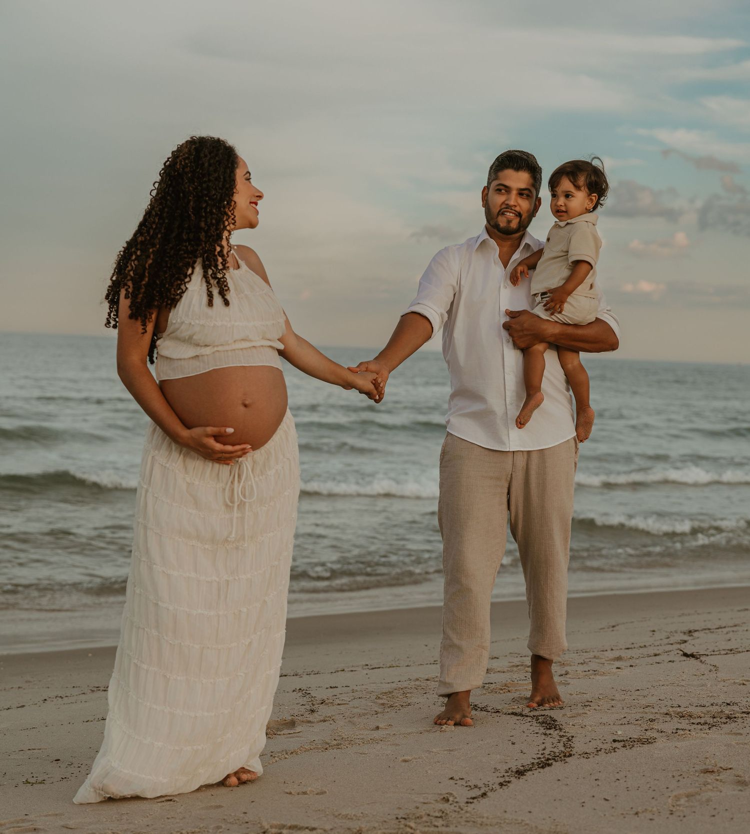 Pregnant woman in white dress holds hands with man carrying toddler on a beach at sunset.
