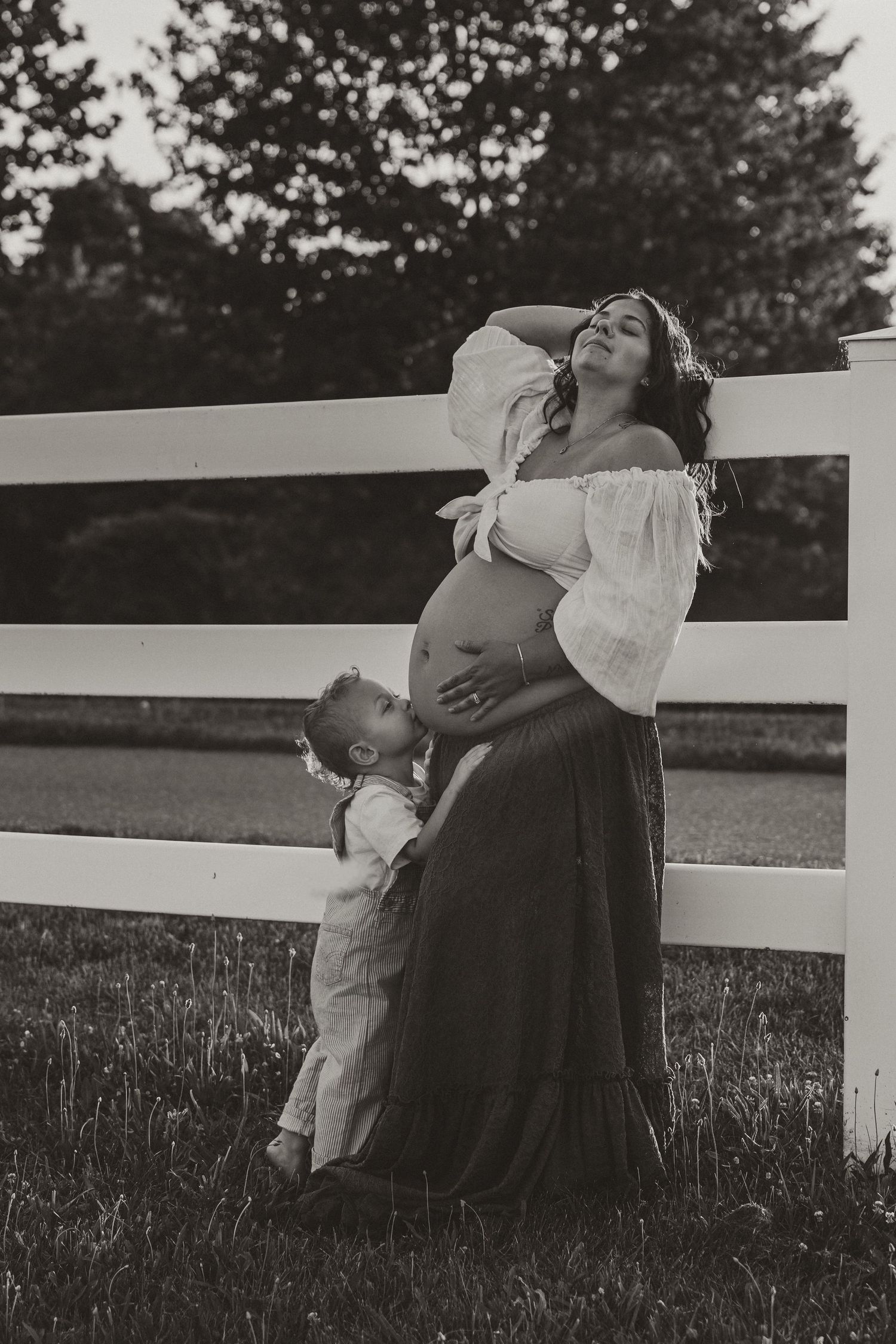 Black and white maternity photo of pregnant woman with toddler kissing her belly by white fence outdoors.