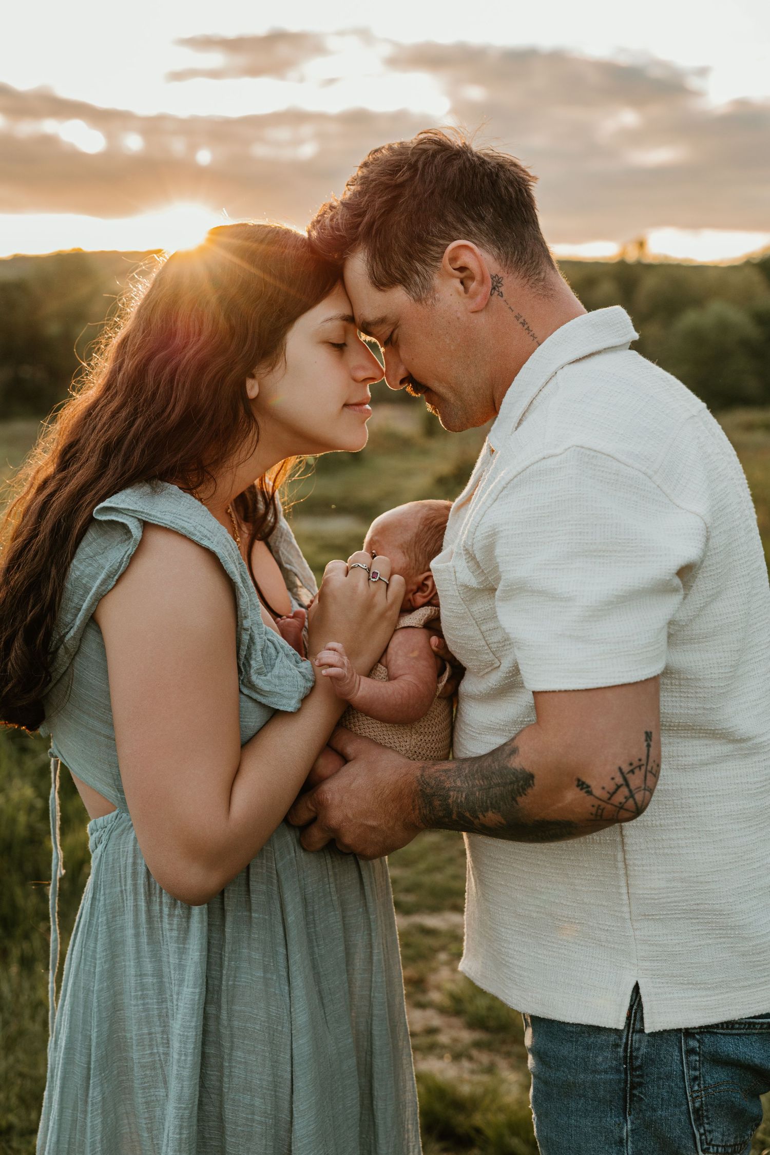 A couple tenderly touches foreheads while holding their newborn baby outdoors at golden sunset.