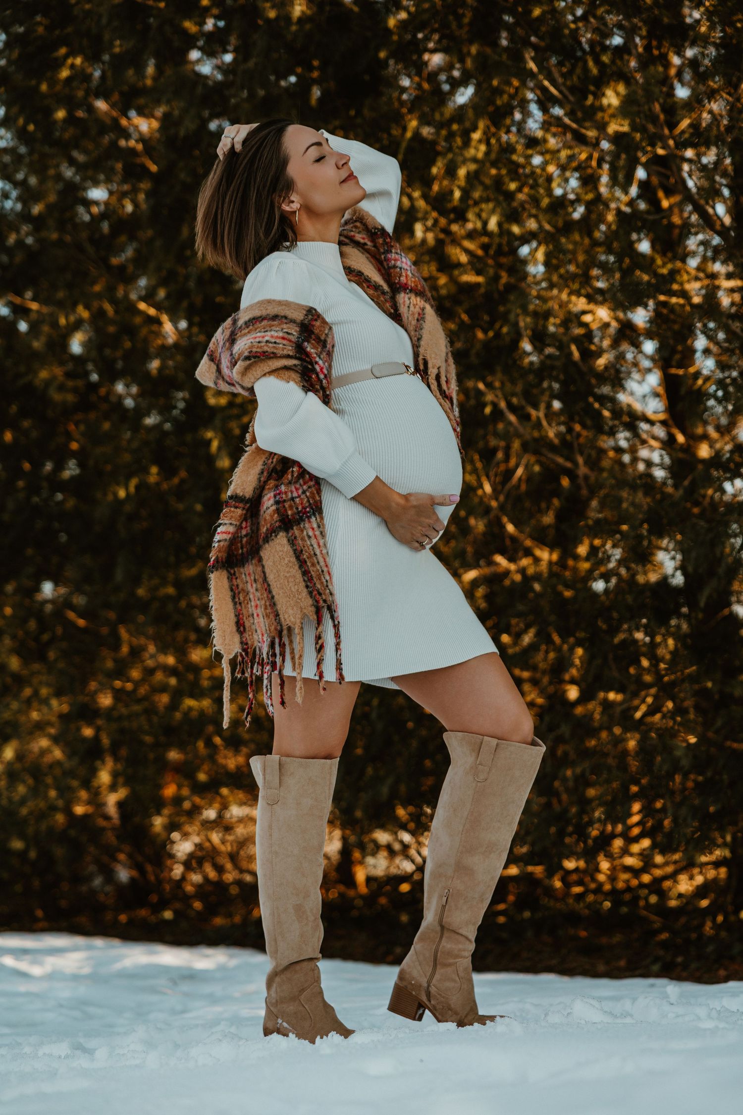 Expectant mother in white dress and plaid shawl poses in snow wearing knee-high boots during golden hour.