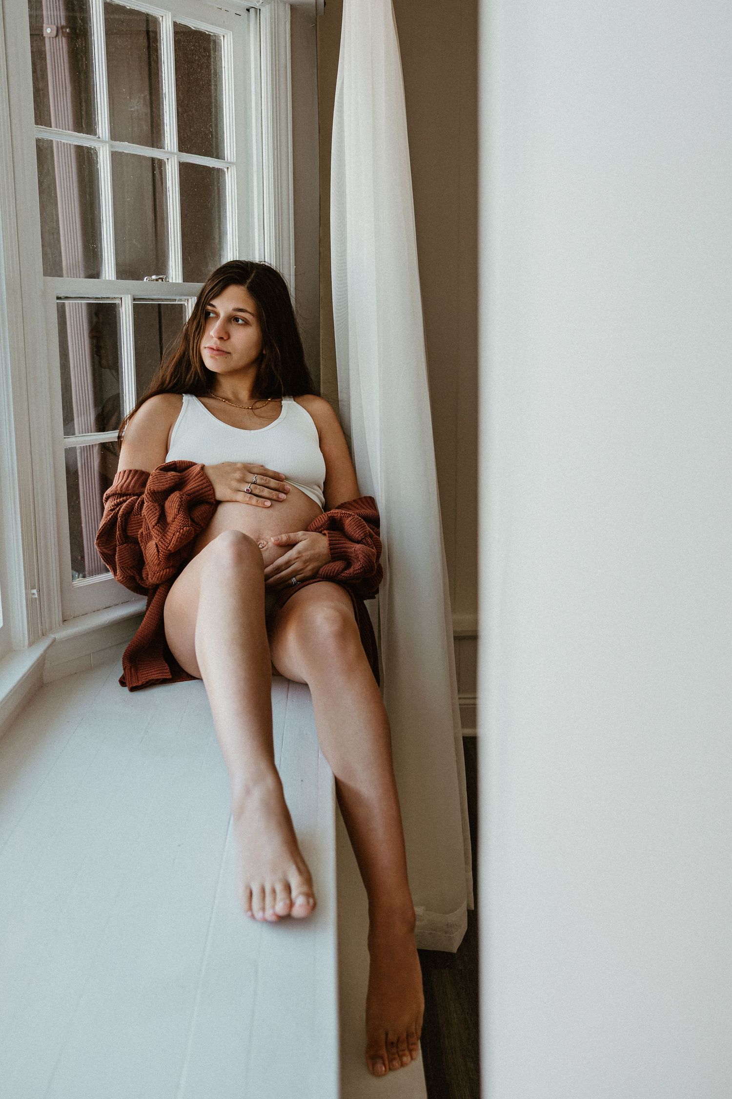 Lifestyle portrait session by a bright window featuring rust-colored outerwear and white tank top.