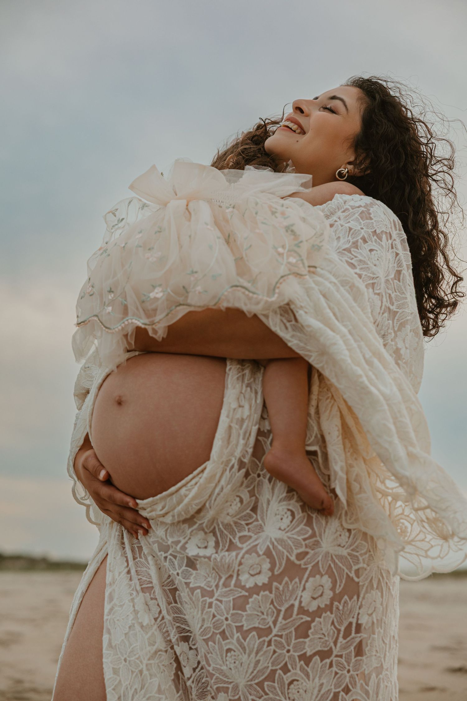 Smiling pregnant woman in elegant lace maternity gown cradling her baby bump on a beach.