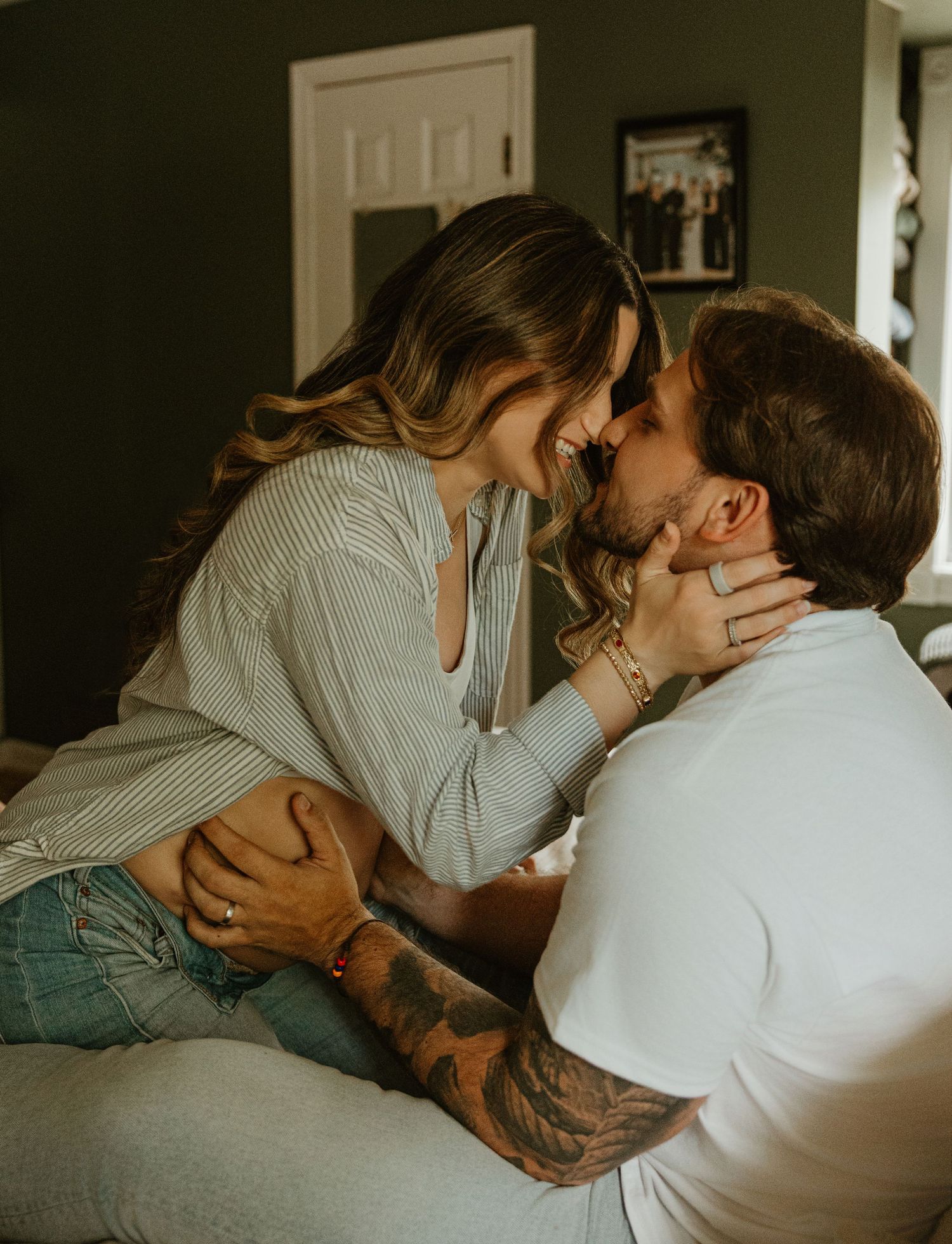 A couple sharing an intimate moment on a couch, smiling and about to kiss, with the man showing tattooed arms.