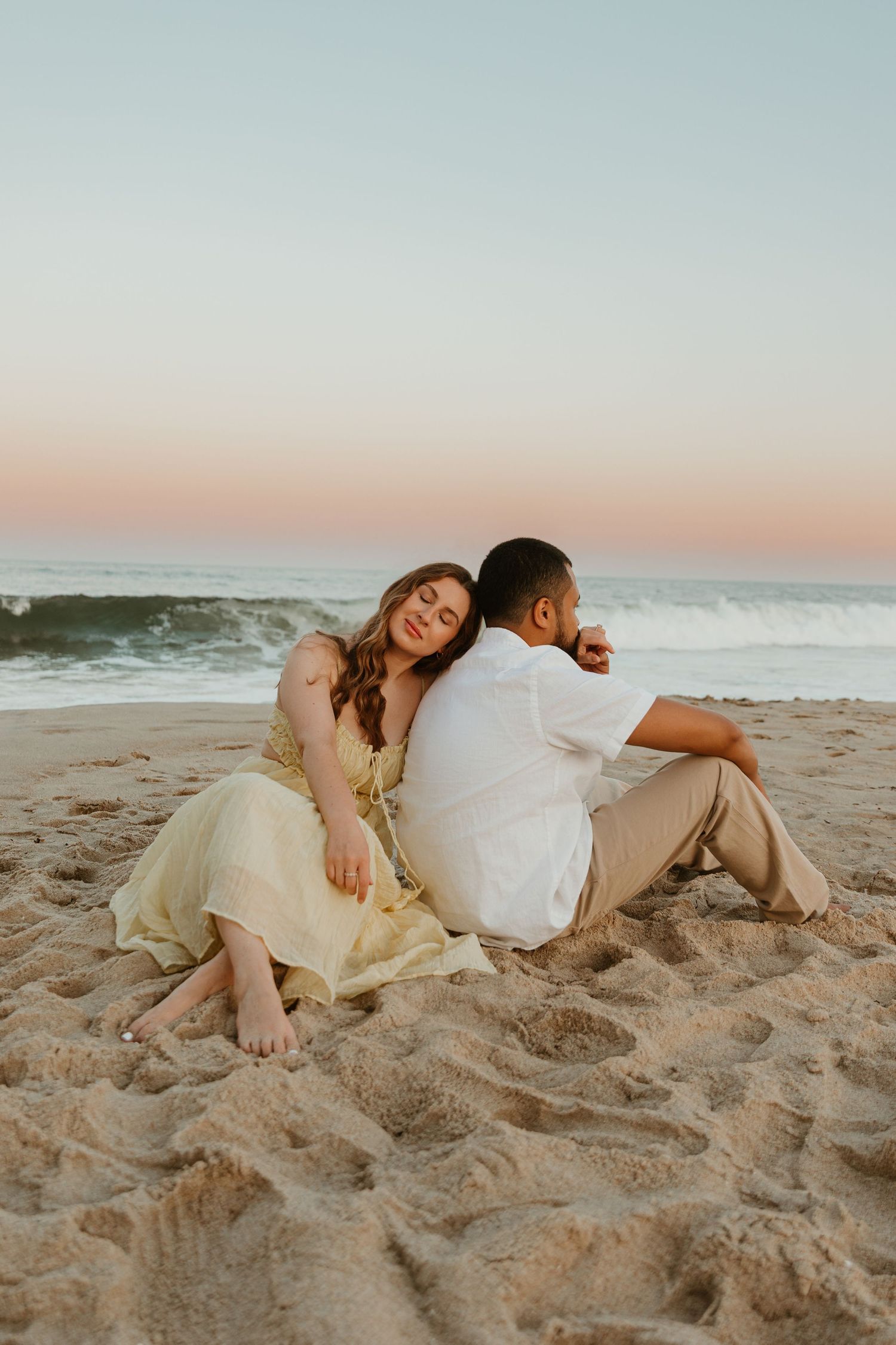 A couple sits back-to-back on a sandy beach at sunset with ocean waves in the background.