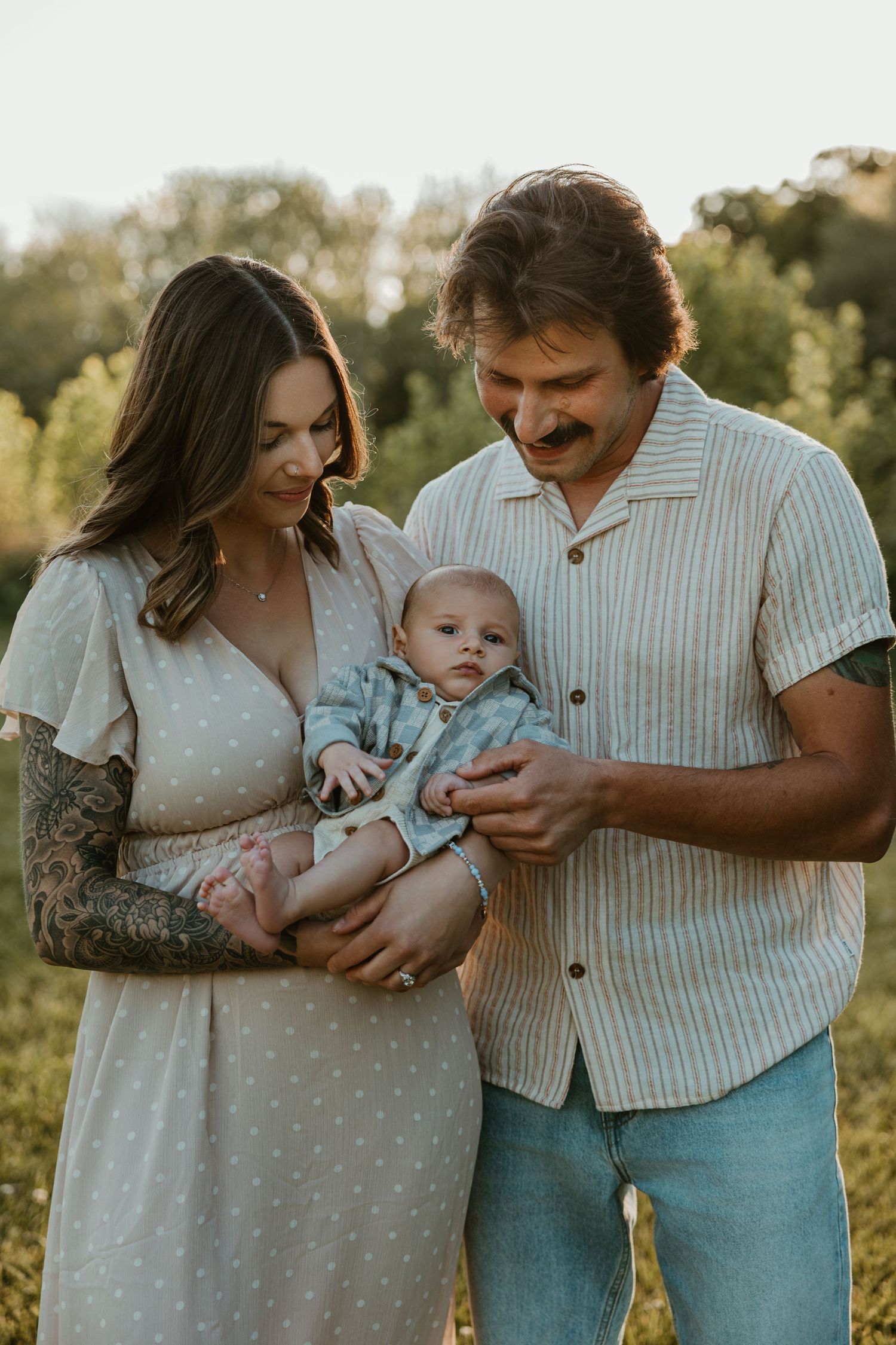 A young couple with tattoos lovingly holds their newborn baby outdoors in a sunlit green field.