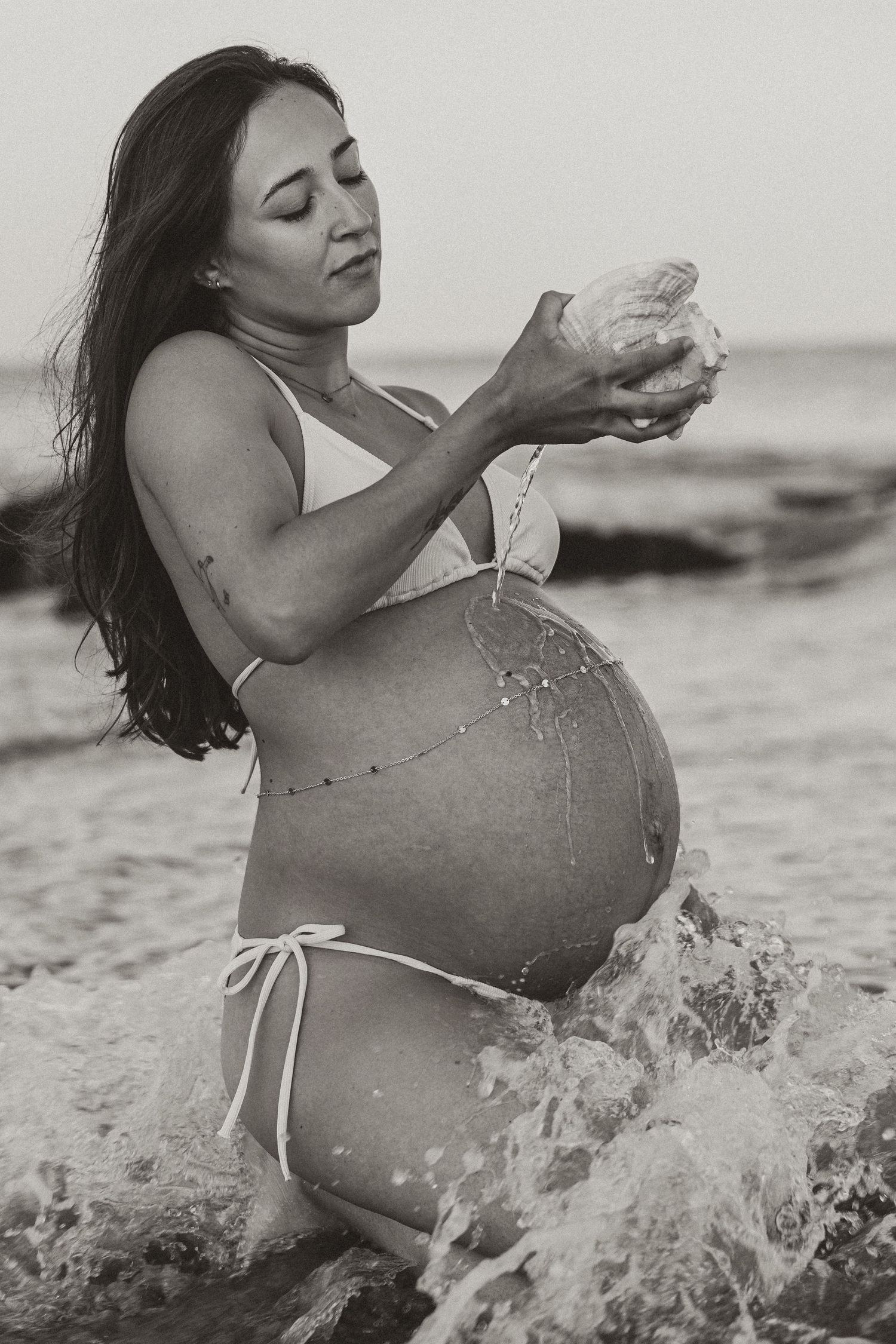 Black and white photo of a pregnant woman in a bikini holding a coconut while standing in ocean waves.