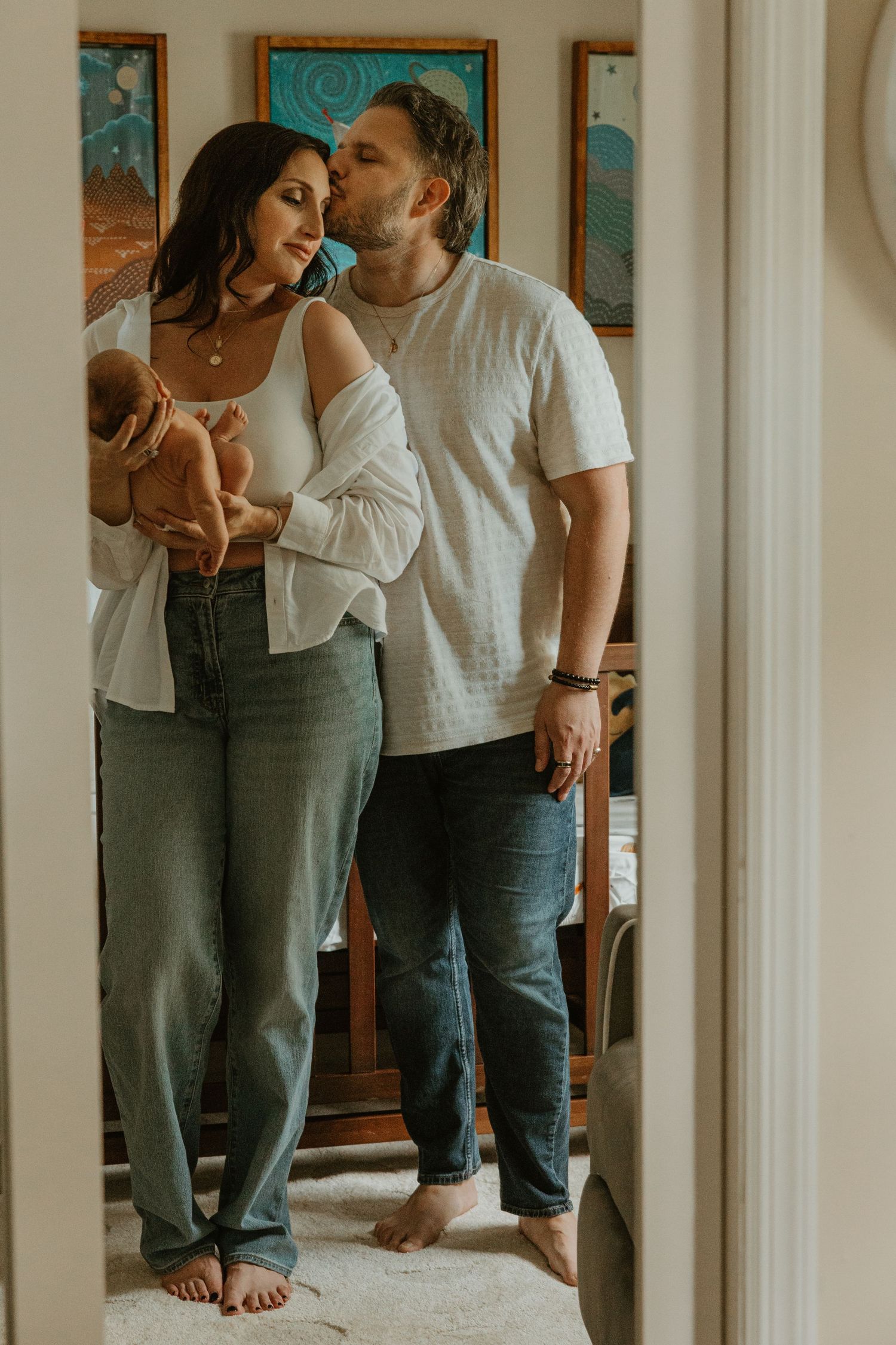 A couple in casual white tops and jeans share a tender kiss while holding their newborn baby at home.