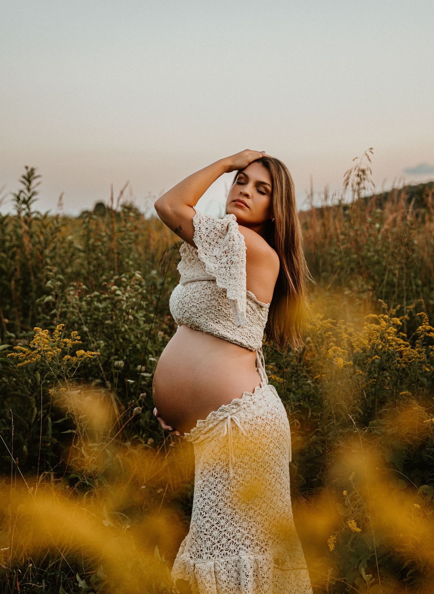 Artistic maternity photo in wildflower field with golden sunlight and lace dress.