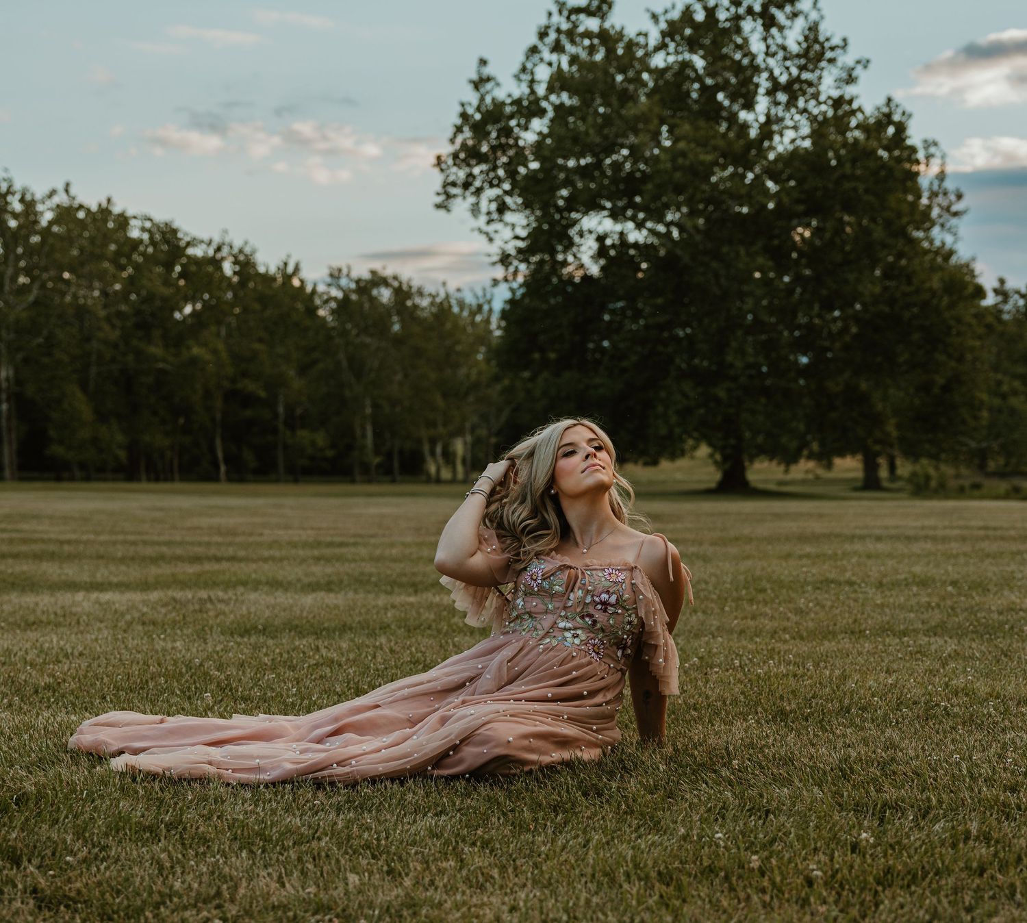A woman in a flowing pink beaded gown sits gracefully on lush green grass with trees in the background.