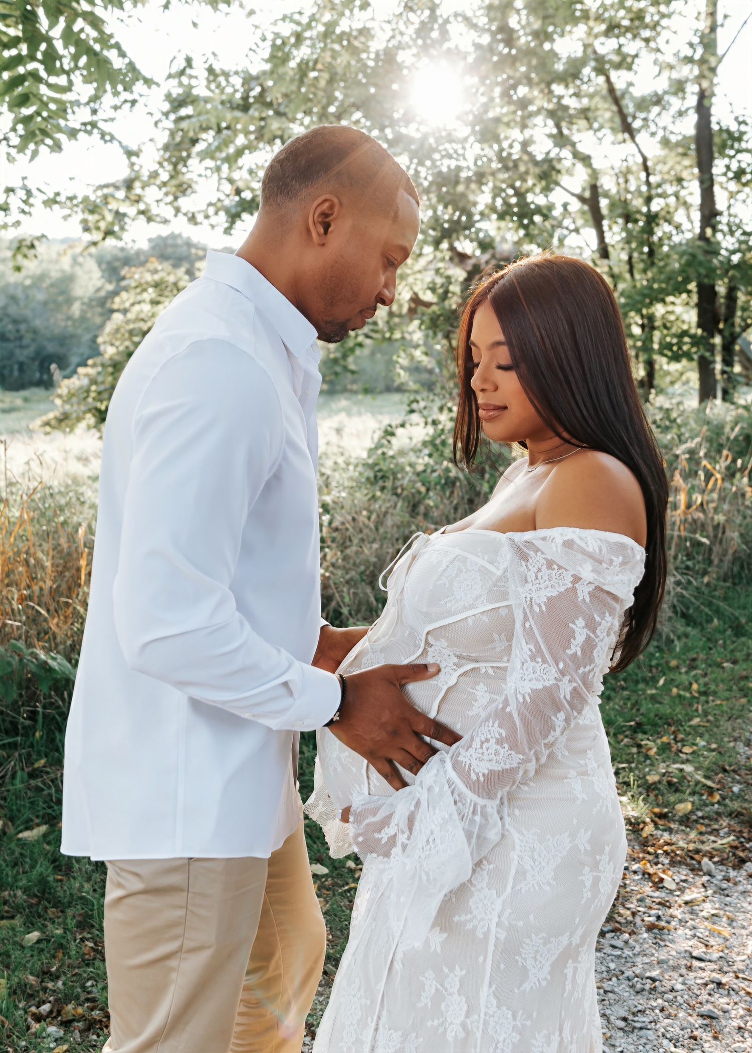 A couple shares a tender moment in a sunlit field, celebrating an expected arrival.