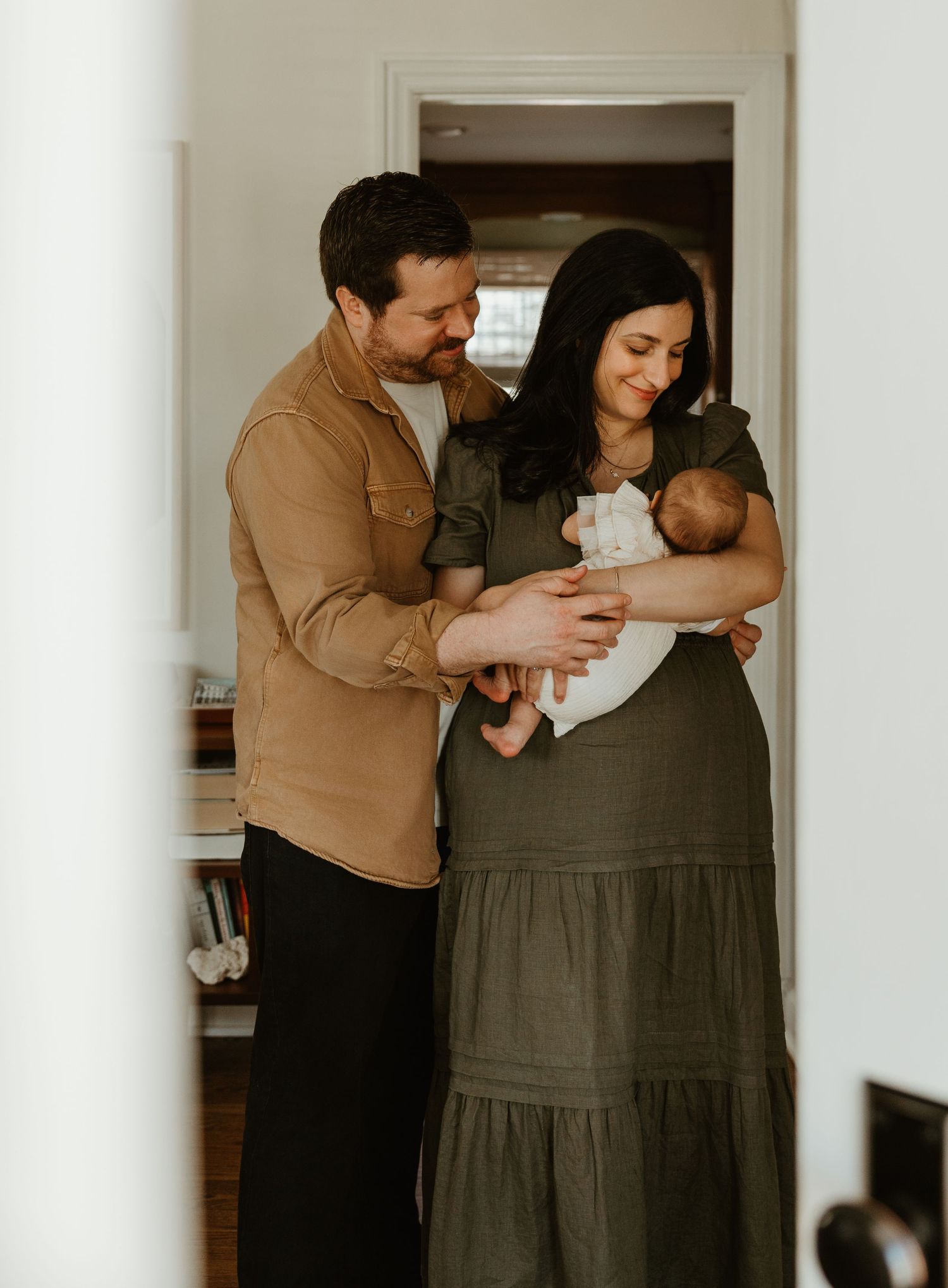A couple in a doorway lovingly gazes at their newborn baby, the mother in olive dress, father in tan shirt.