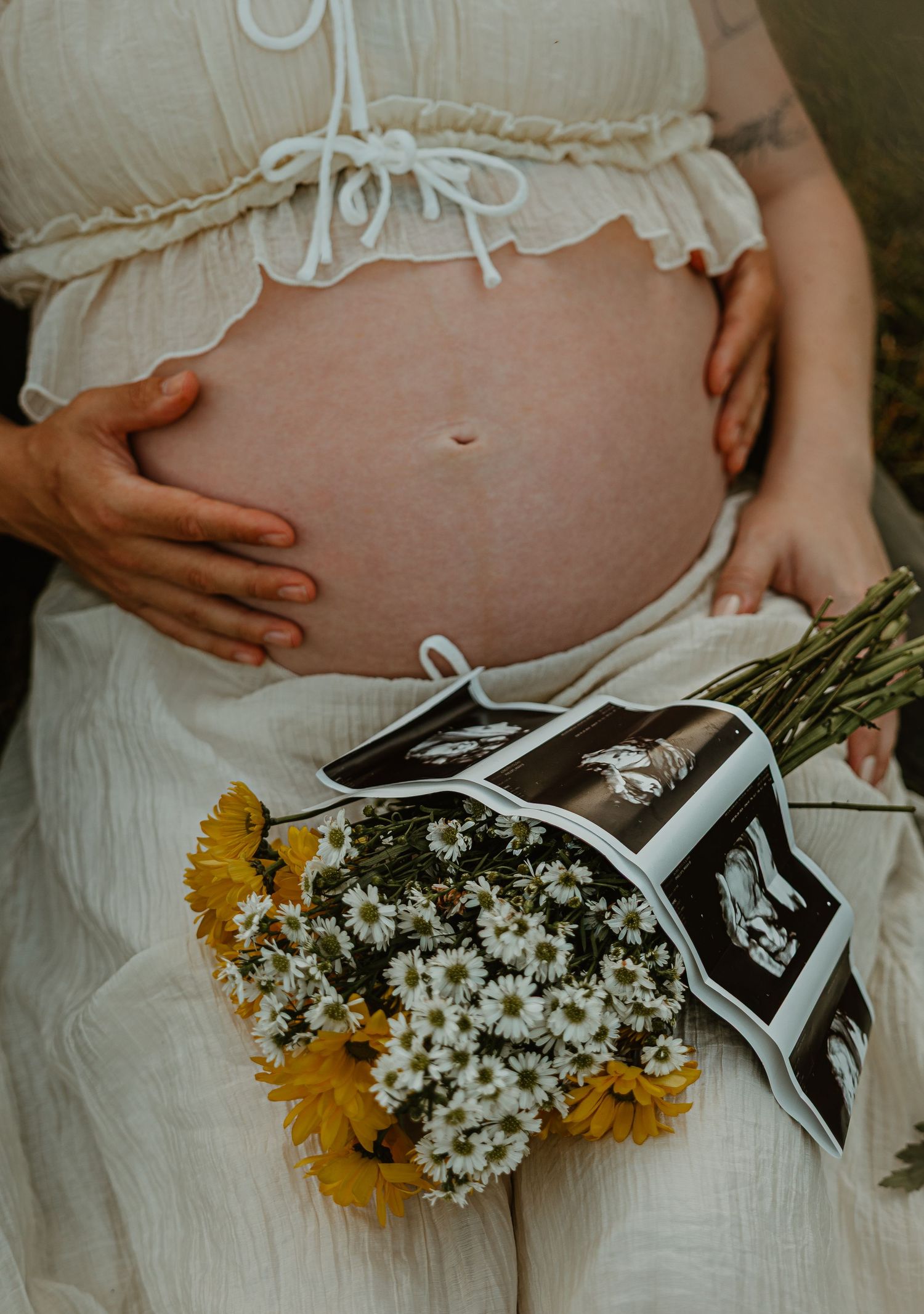 Pregnant woman in white dress holding baby ultrasound photos with daisy bouquet during maternity photoshoot.